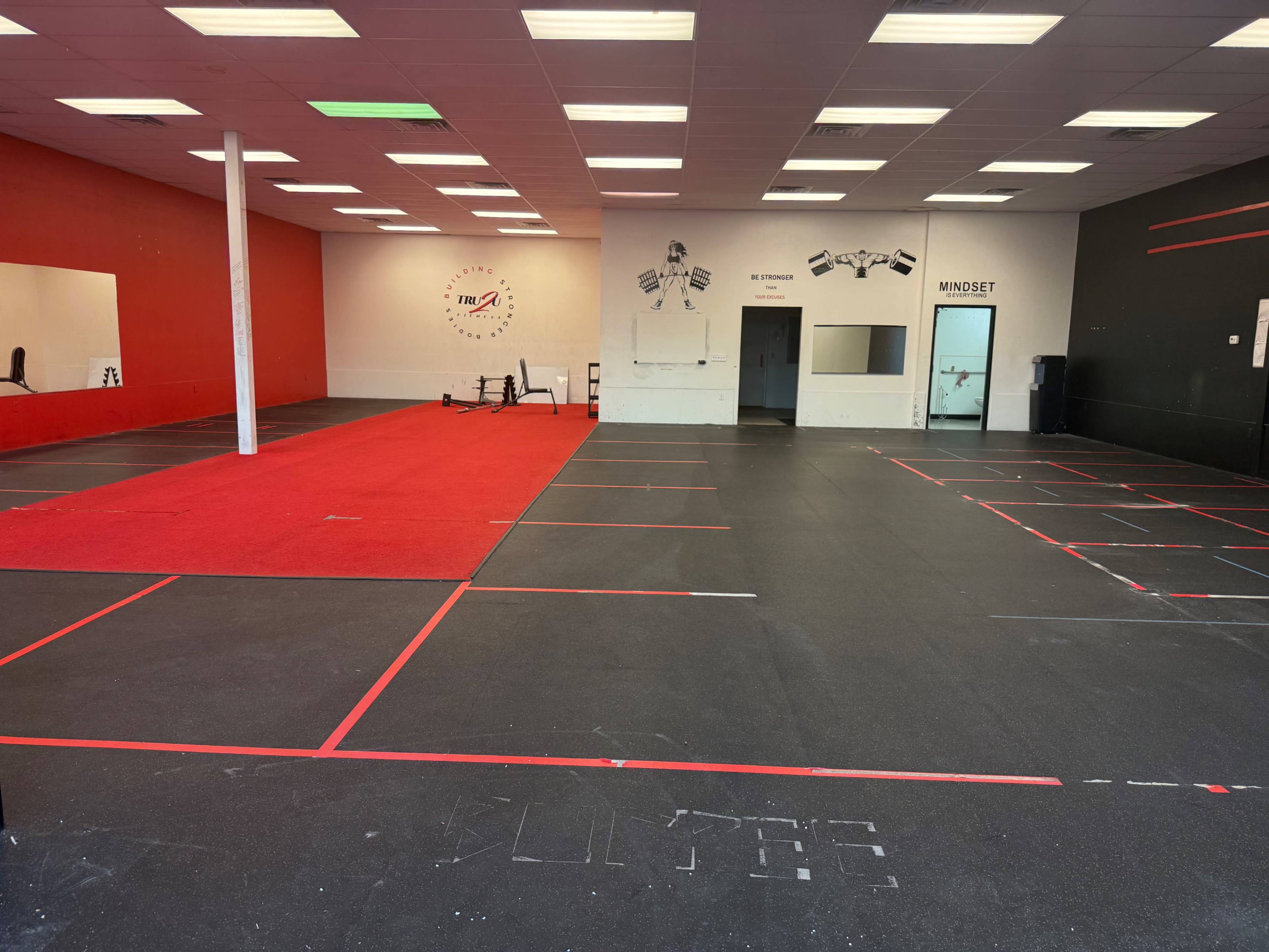 The image features a spacious gym interior with black rubber flooring, red-marked sections, and walls painted in red and white, along with some exercise equipment visible in the background.