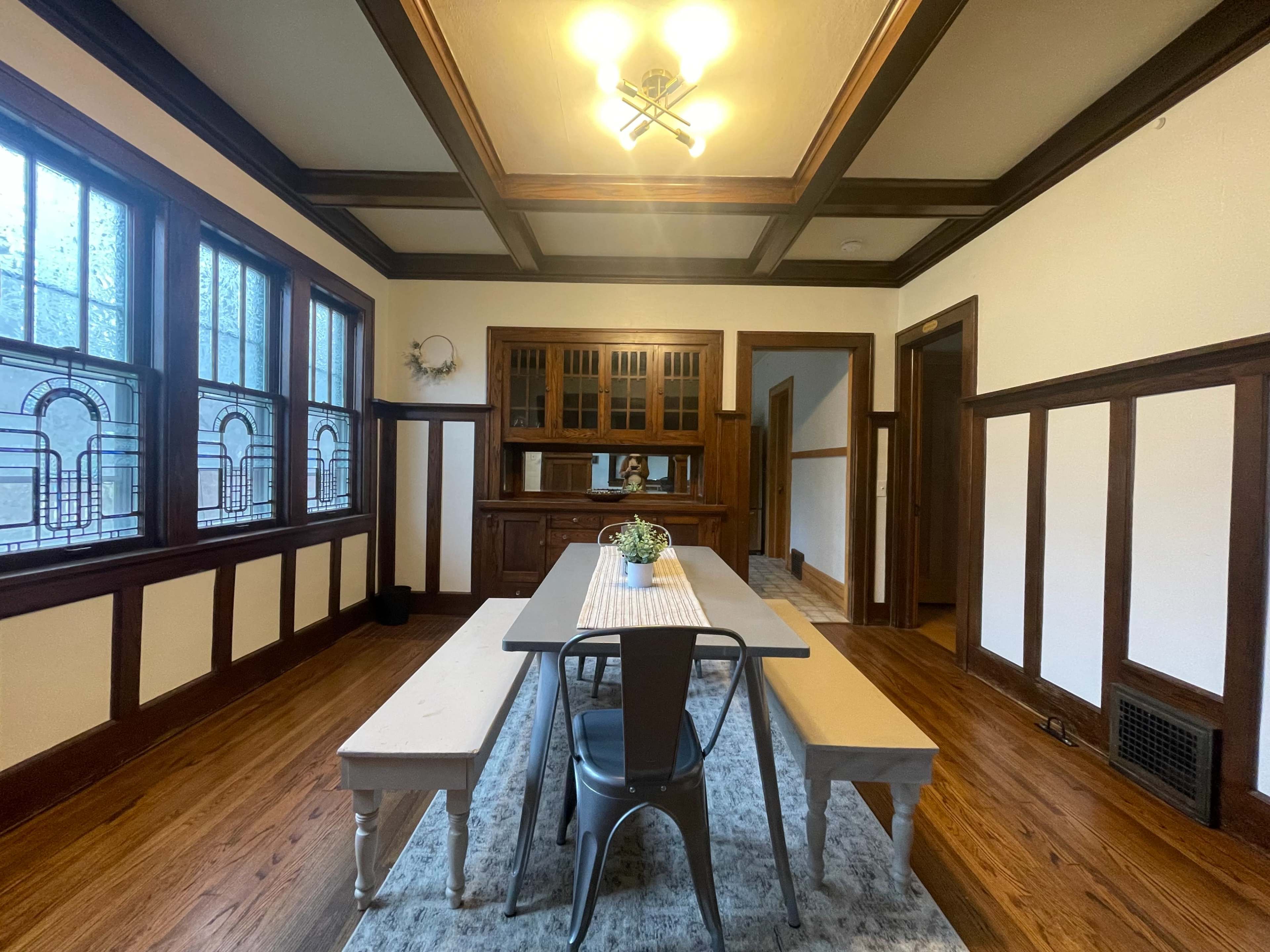 A dining room with a long table and benches, framed by wooden paneling and large windows.