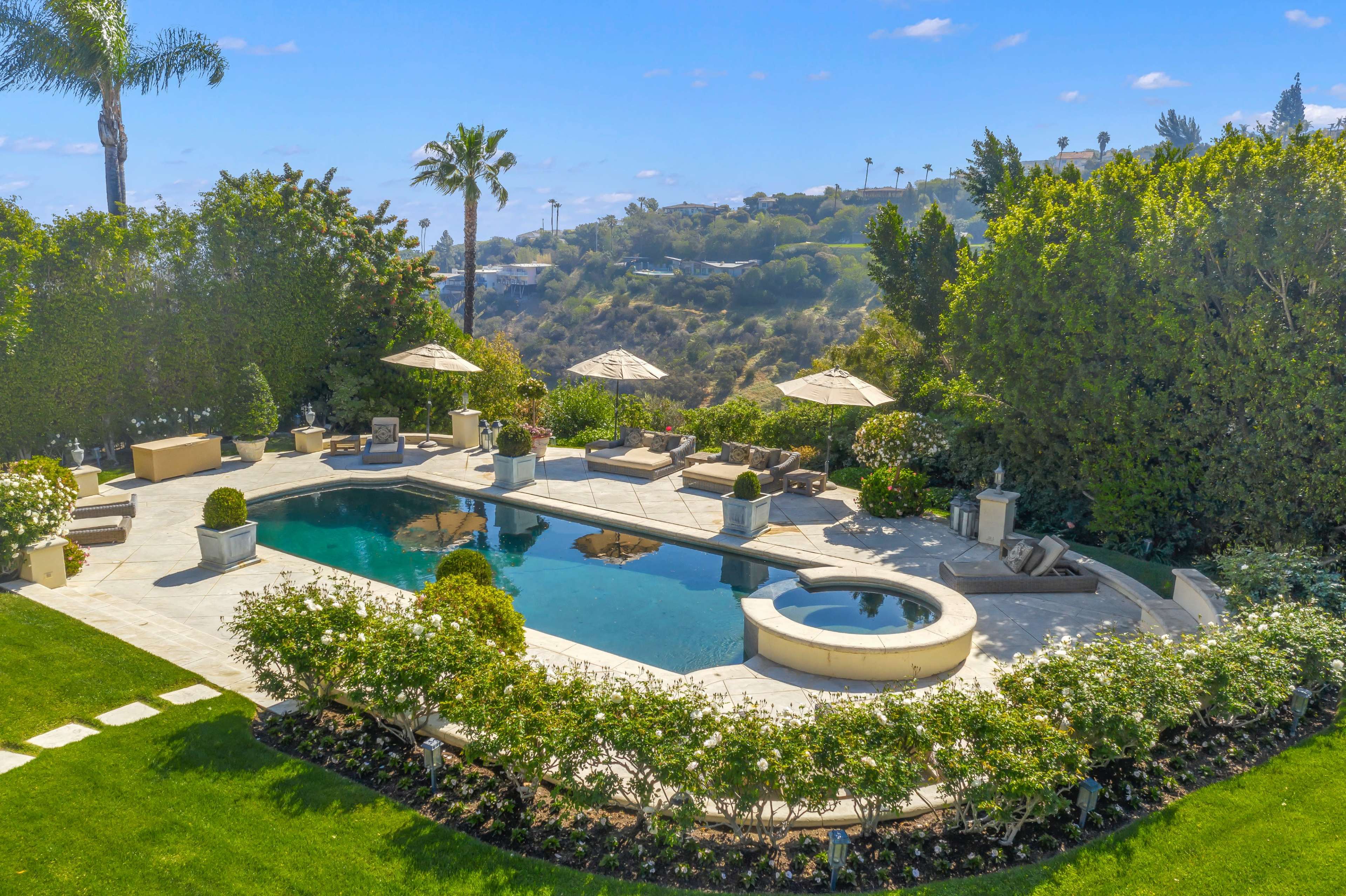 The image shows a landscaped backyard featuring a swimming pool surrounded by lounge chairs and umbrellas, with lush greenery and well-maintained flower beds.