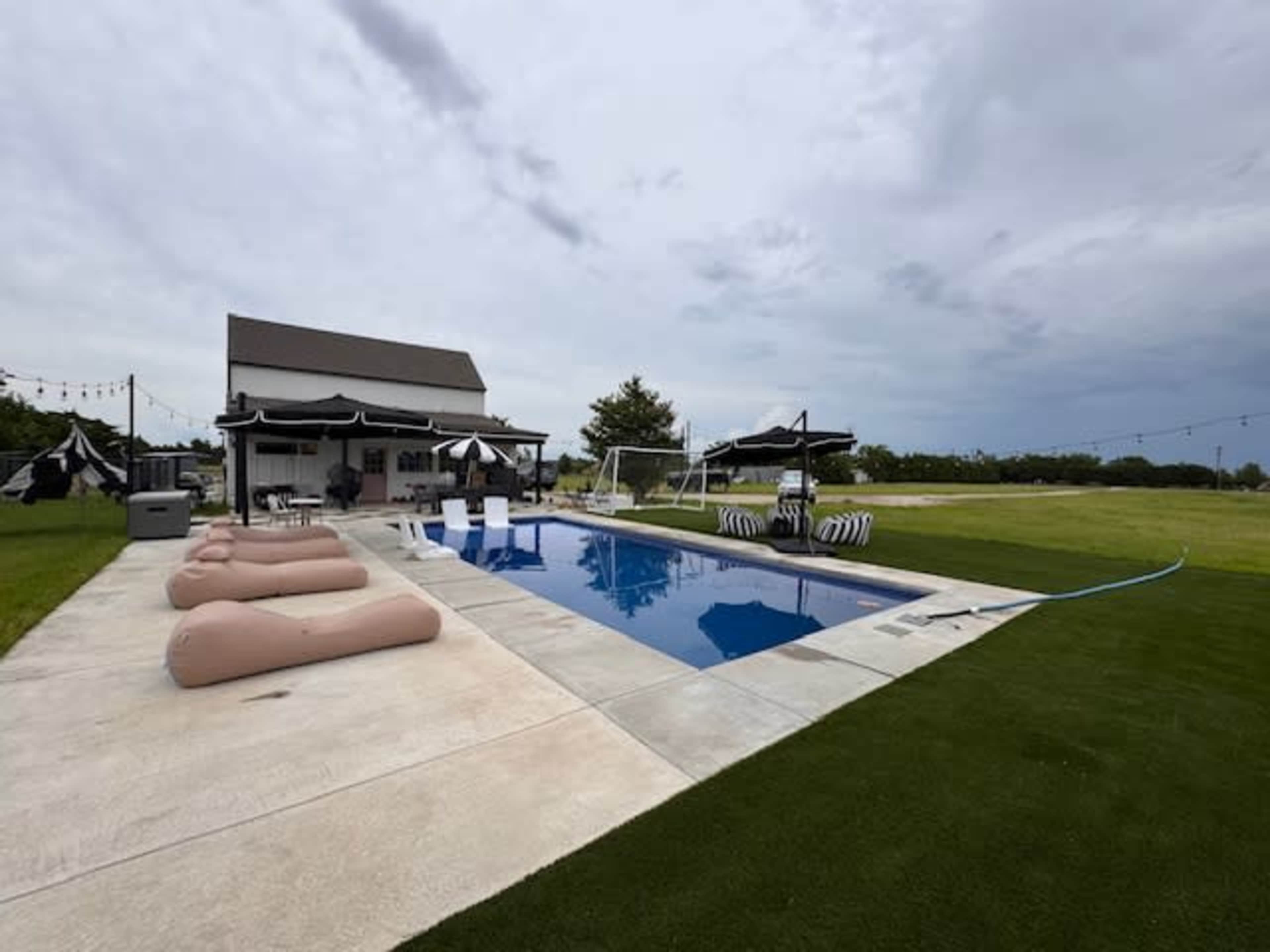 The scene features a swimming pool with lounging chairs and a white house in the background, surrounded by a grassy area under cloudy skies.