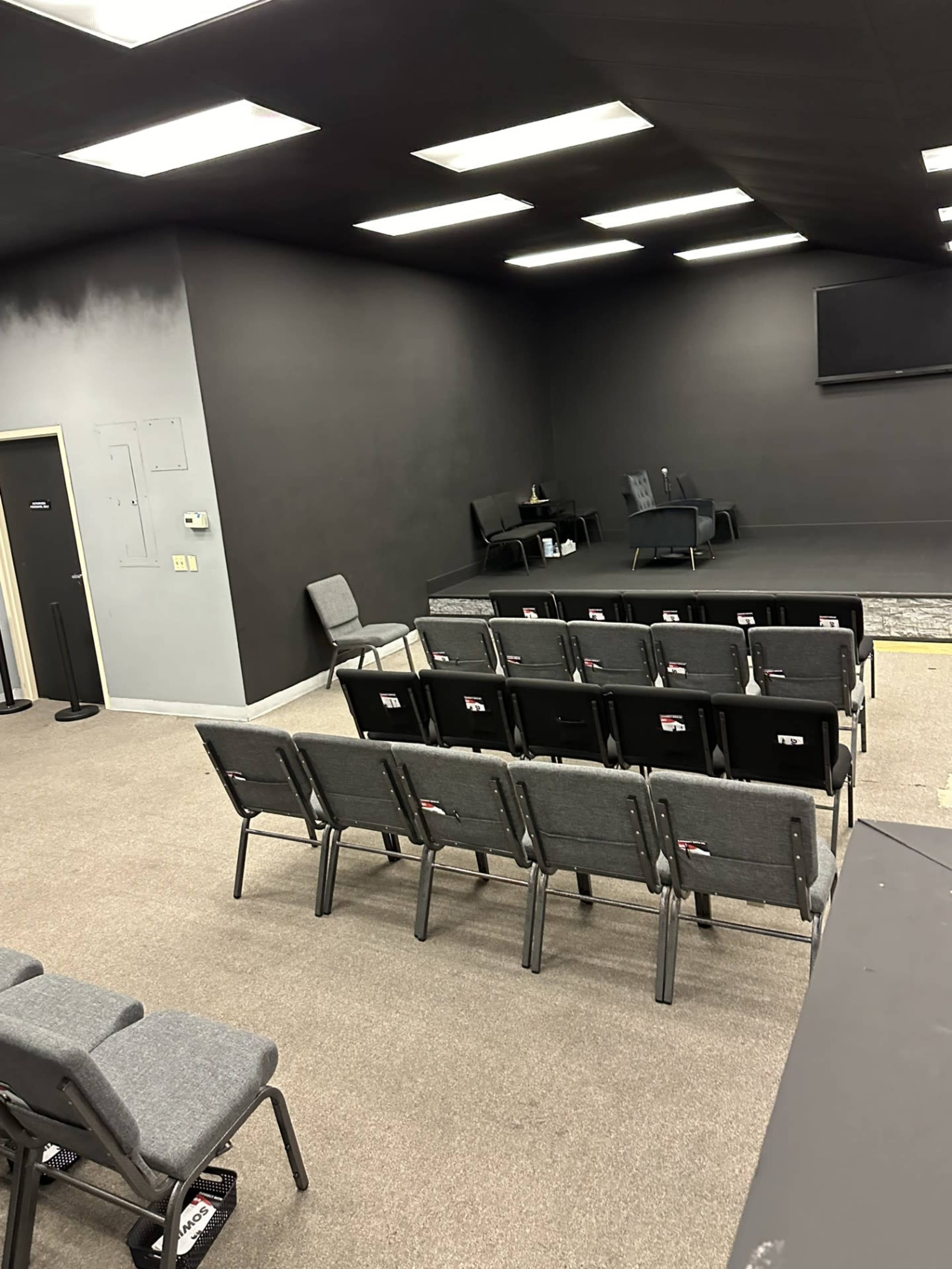 A small auditorium or meeting room with rows of gray chairs facing a stage area, all set against a black wall and a carpeted floor.