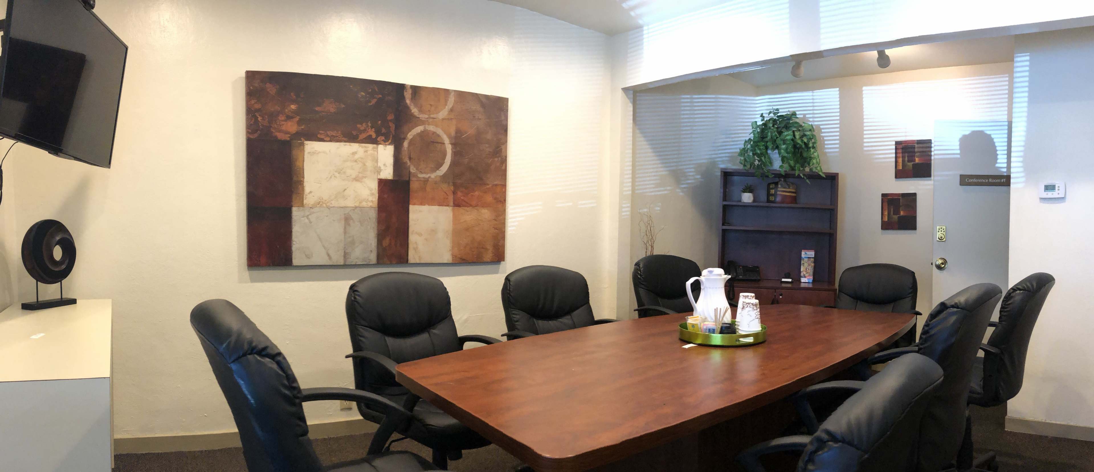 A conference room features a large wooden table surrounded by black chairs, a television, a piece of abstract art on the wall, and a small plant in the corner.
