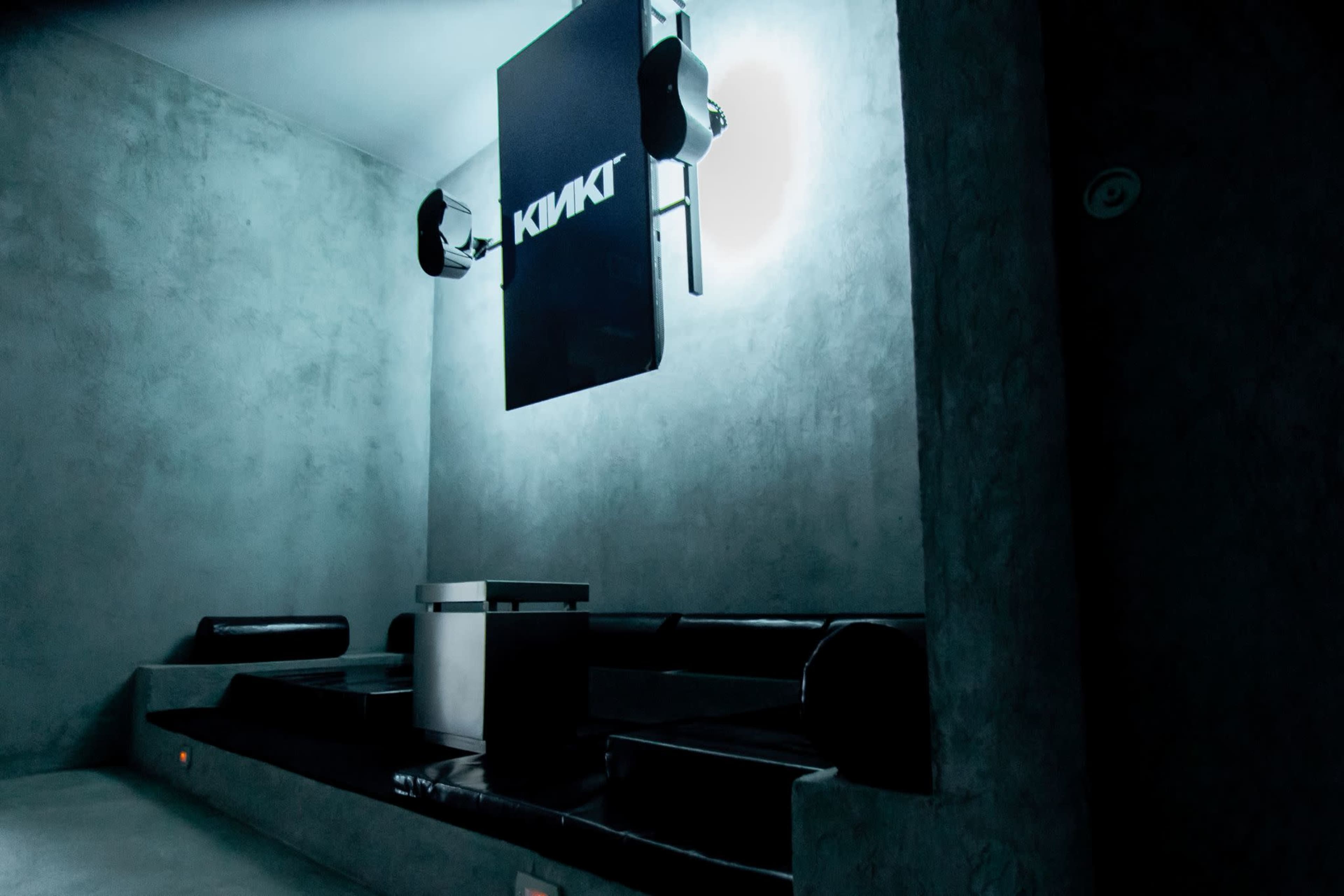 The image shows a minimalistic, dimly lit room with a gray concrete wall and a large black sign that reads "KIKKI."