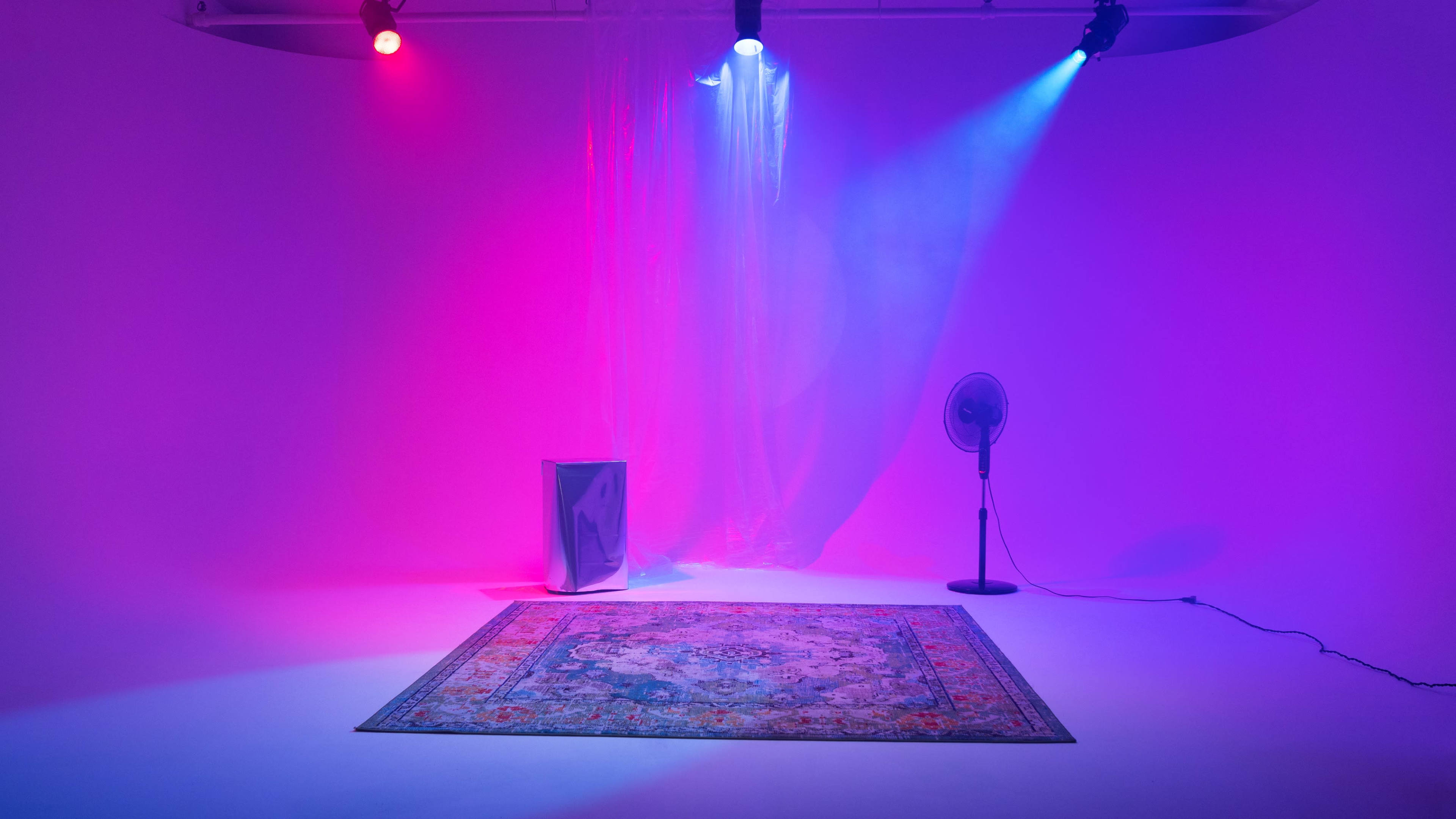 A rectangular rug is positioned on a white floor in an empty studio space illuminated by colorful purple and blue lights, with a fan and a metallic object in the background.