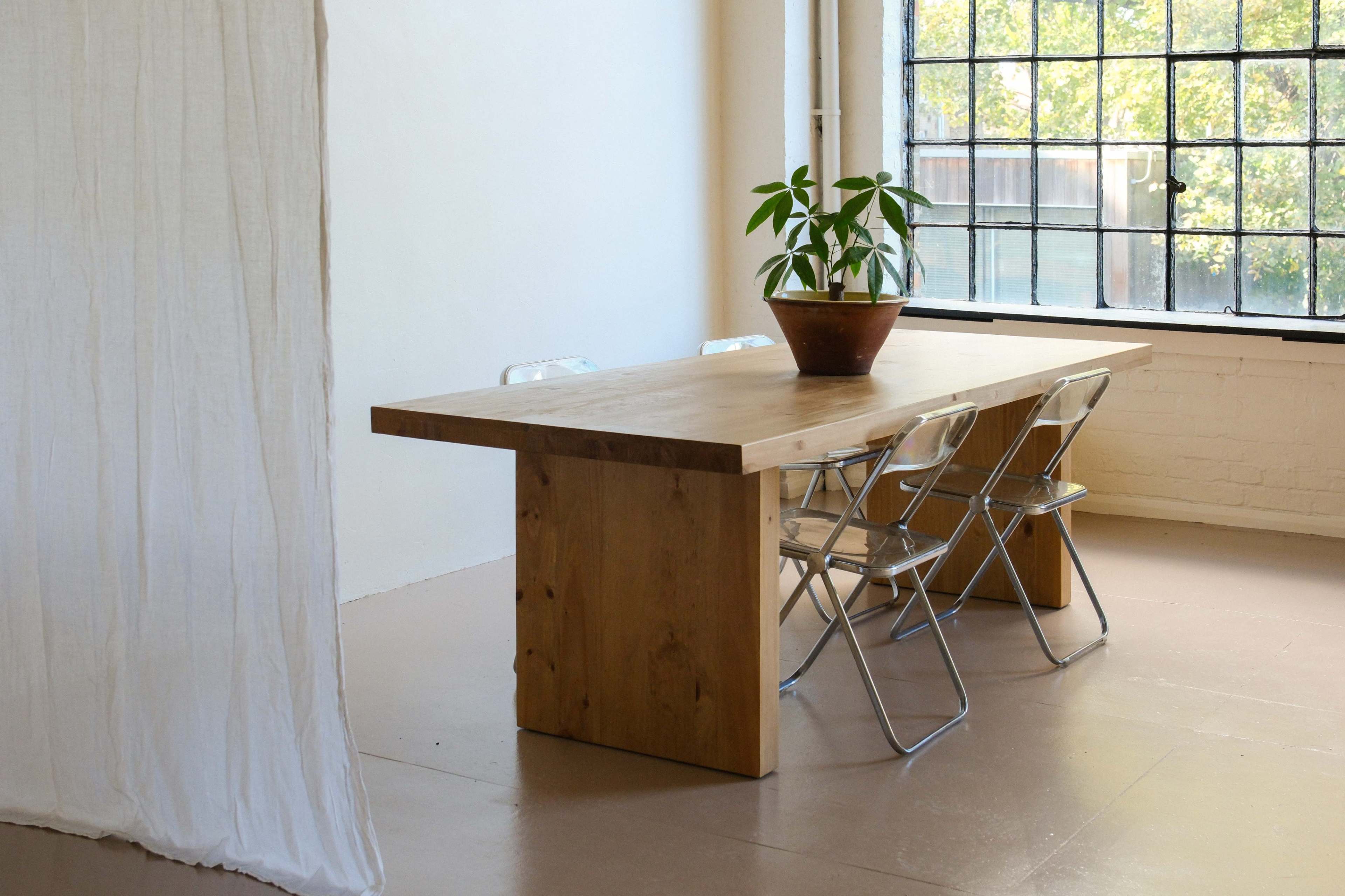 A wooden table with a potted plant sits in a bright room next to a row of metal chairs and a large window.