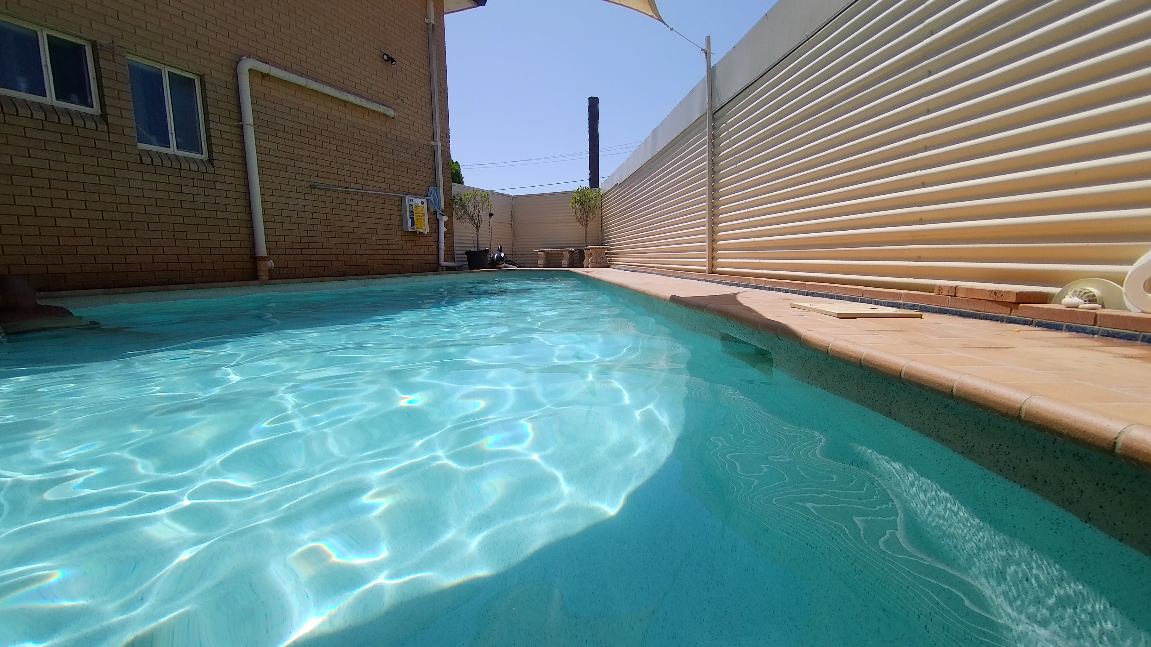 The image shows a clear swimming pool with sunlight reflecting off the water, bordered by a smooth tile edge and a surrounding wall.
