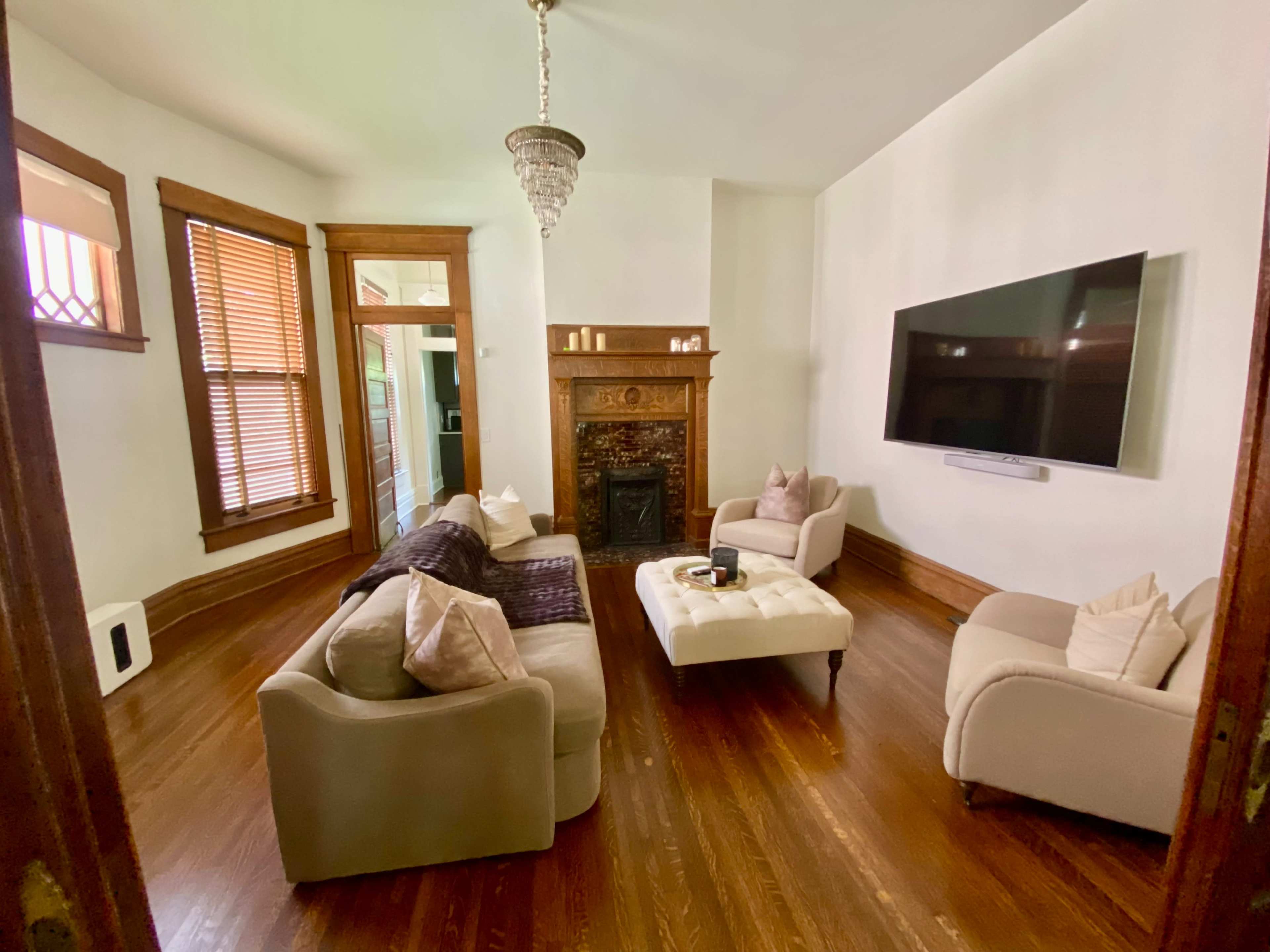 The image shows a living room with light-colored furniture, hardwood floors, and a large flat-screen TV mounted on the wall next to a decorative fireplace.