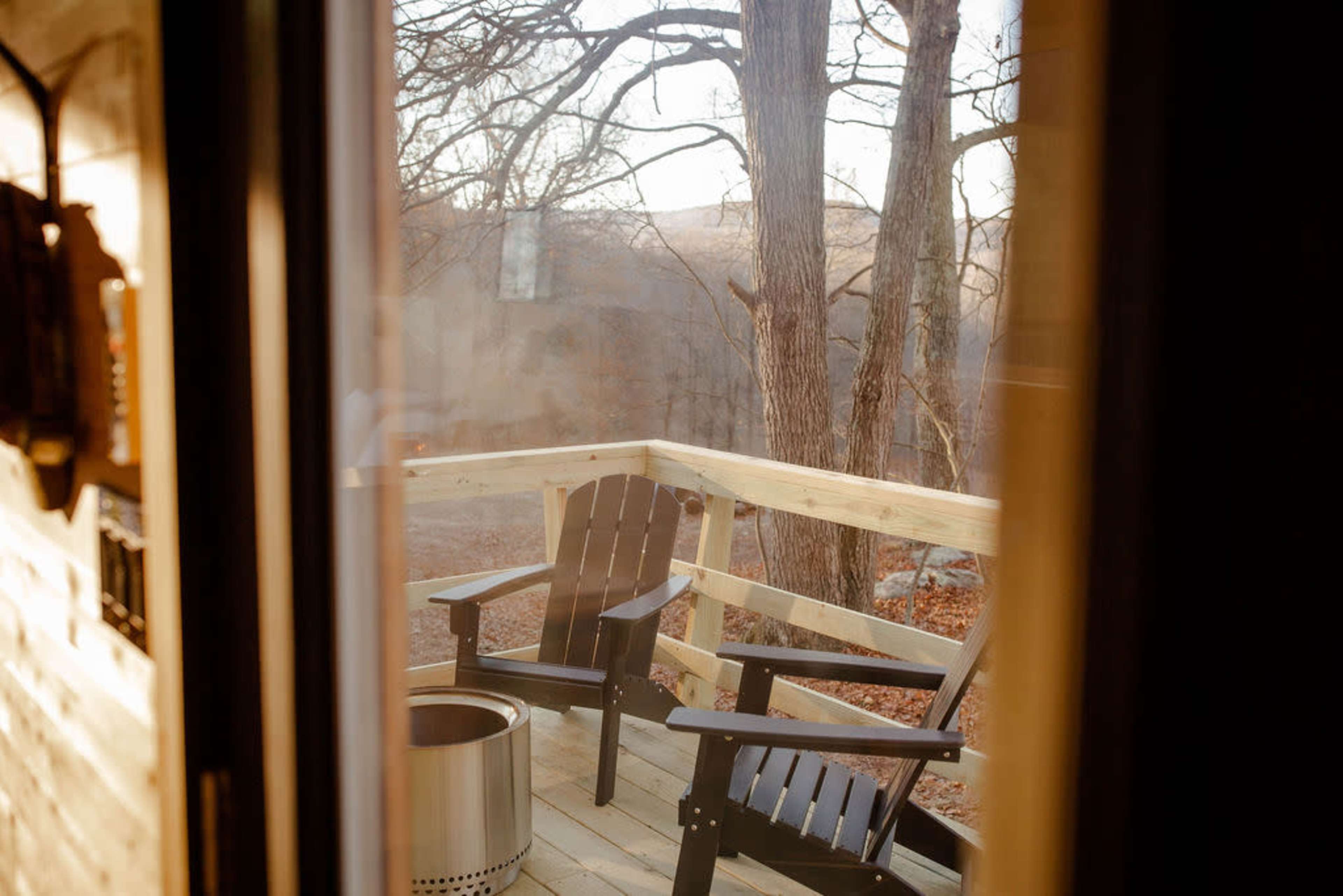 A wooden deck with two black Adirondack chairs and a fire pit, surrounded by trees in the background.