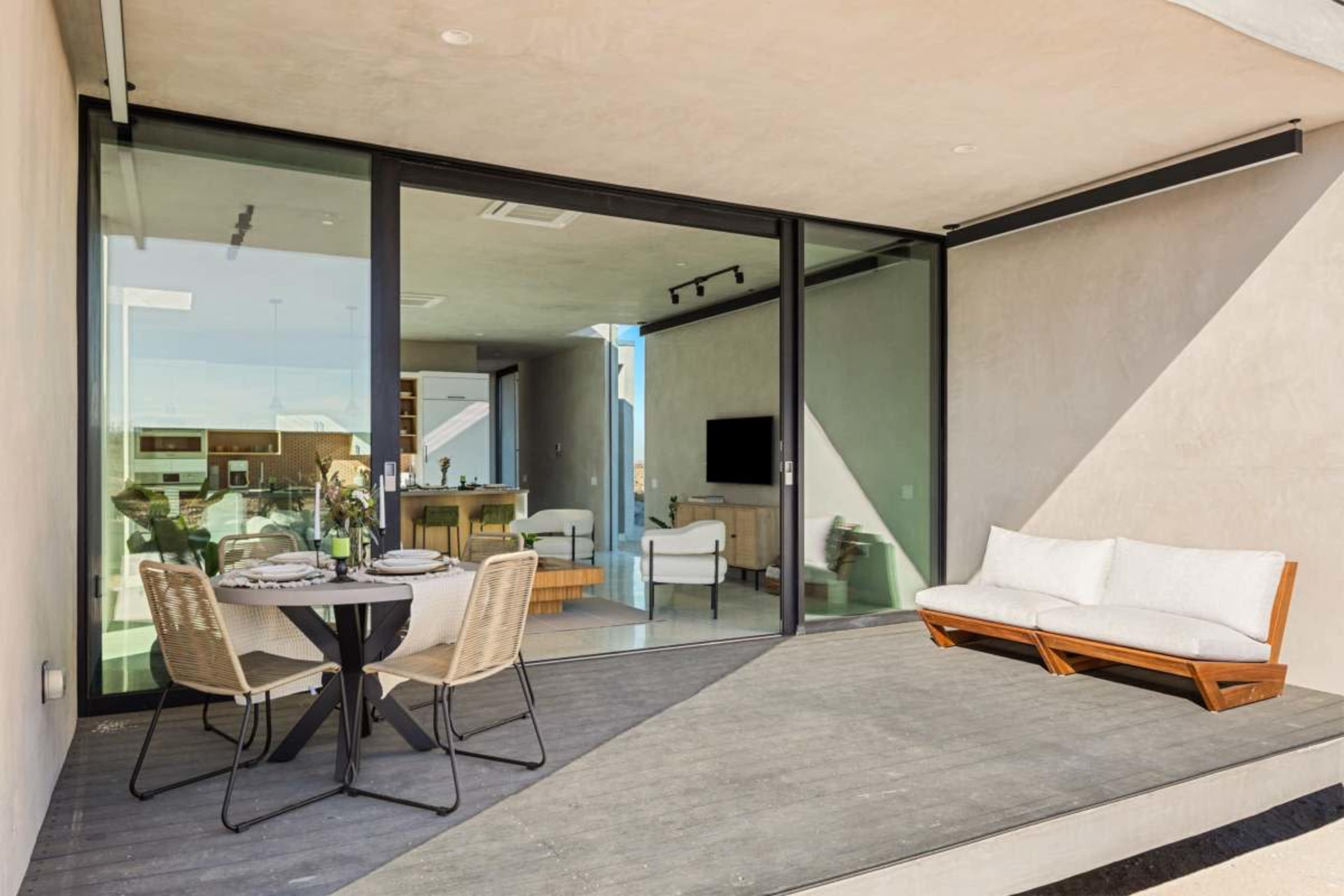 A modern patio features a small dining table with chairs and a sofa, enclosed by large glass sliding doors.