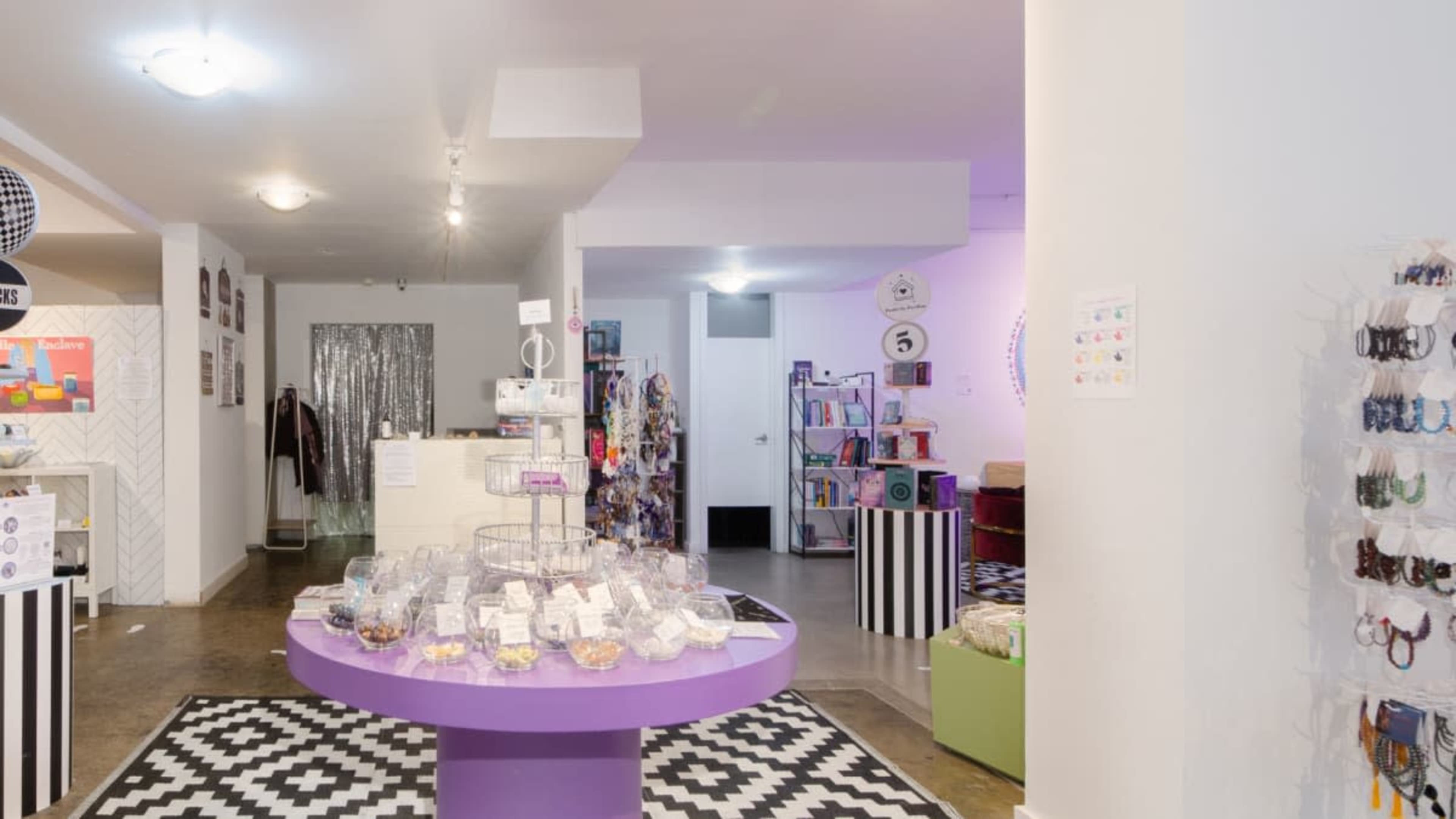 The image shows a brightly lit retail space with a circular purple display table surrounded by various merchandise, including accessories and clothing, against a patterned floor.