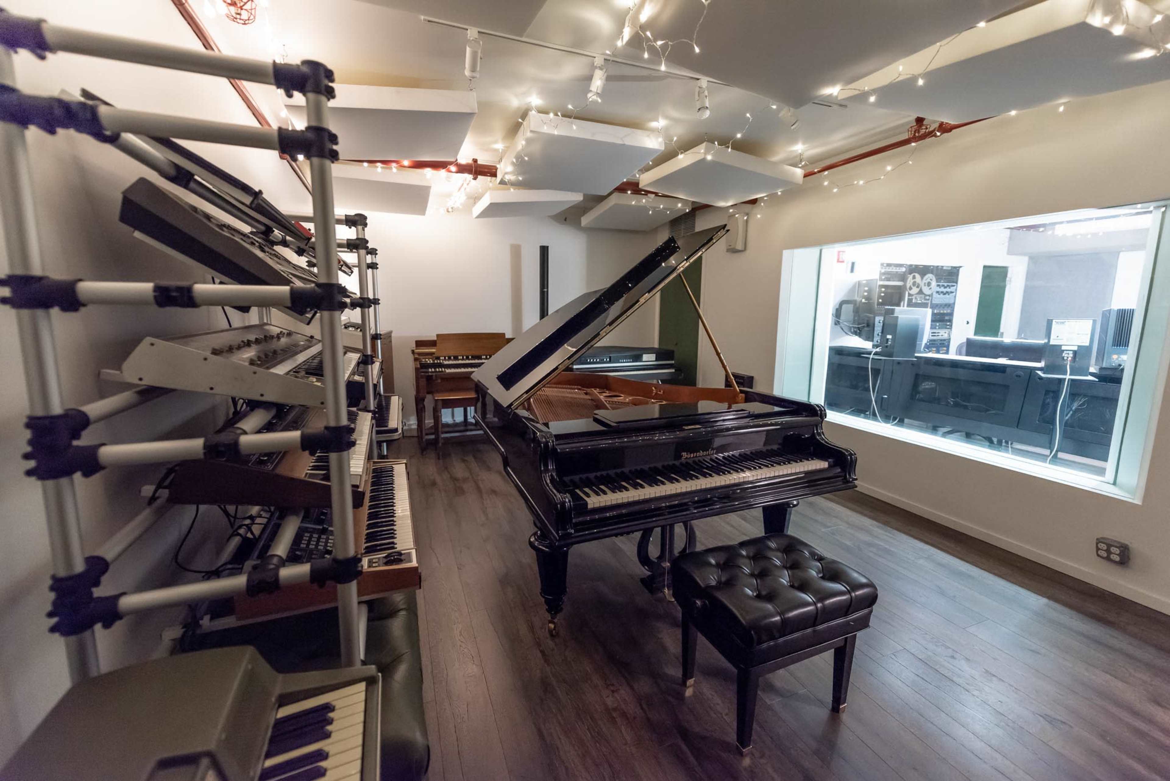 A music studio featuring a grand piano, various electronic instruments on racks, and a glass-walled control room in the background.