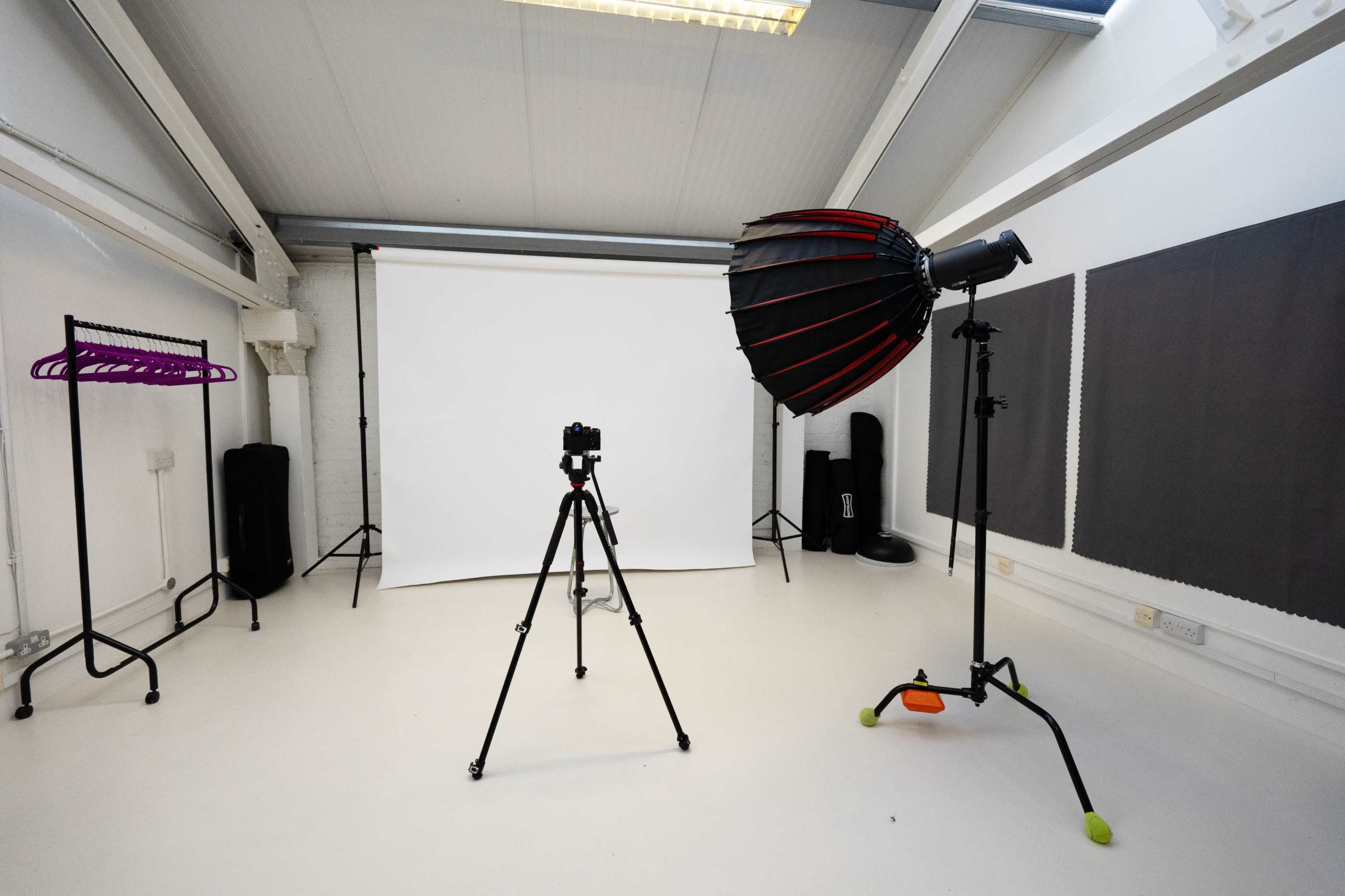 The image shows a photography studio with a camera on a tripod, an umbrella light, a white backdrop, and various props arranged around the space.