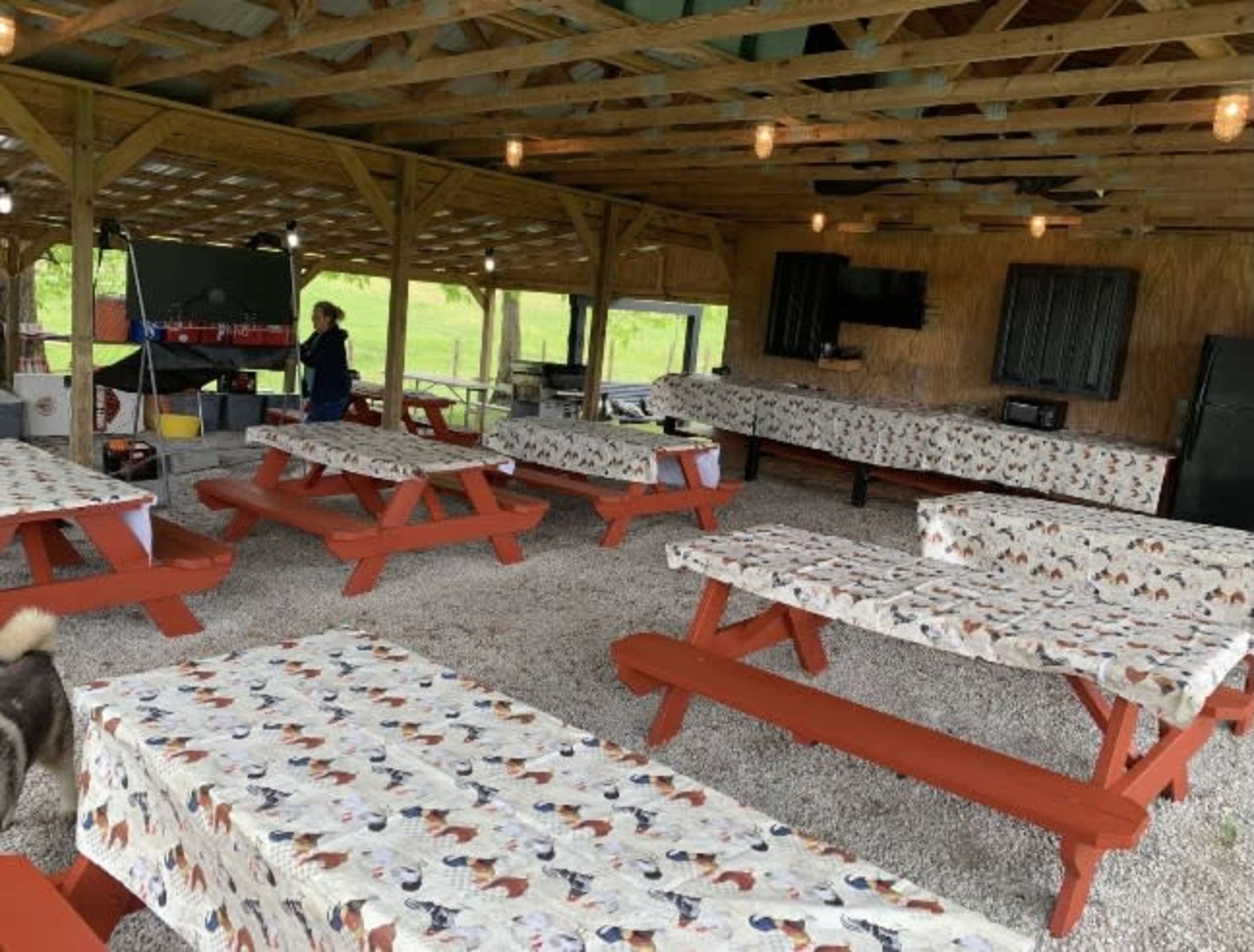 The image shows a covered outdoor dining area with red picnic tables covered in patterned tablecloths and a kitchen setup in the back.
