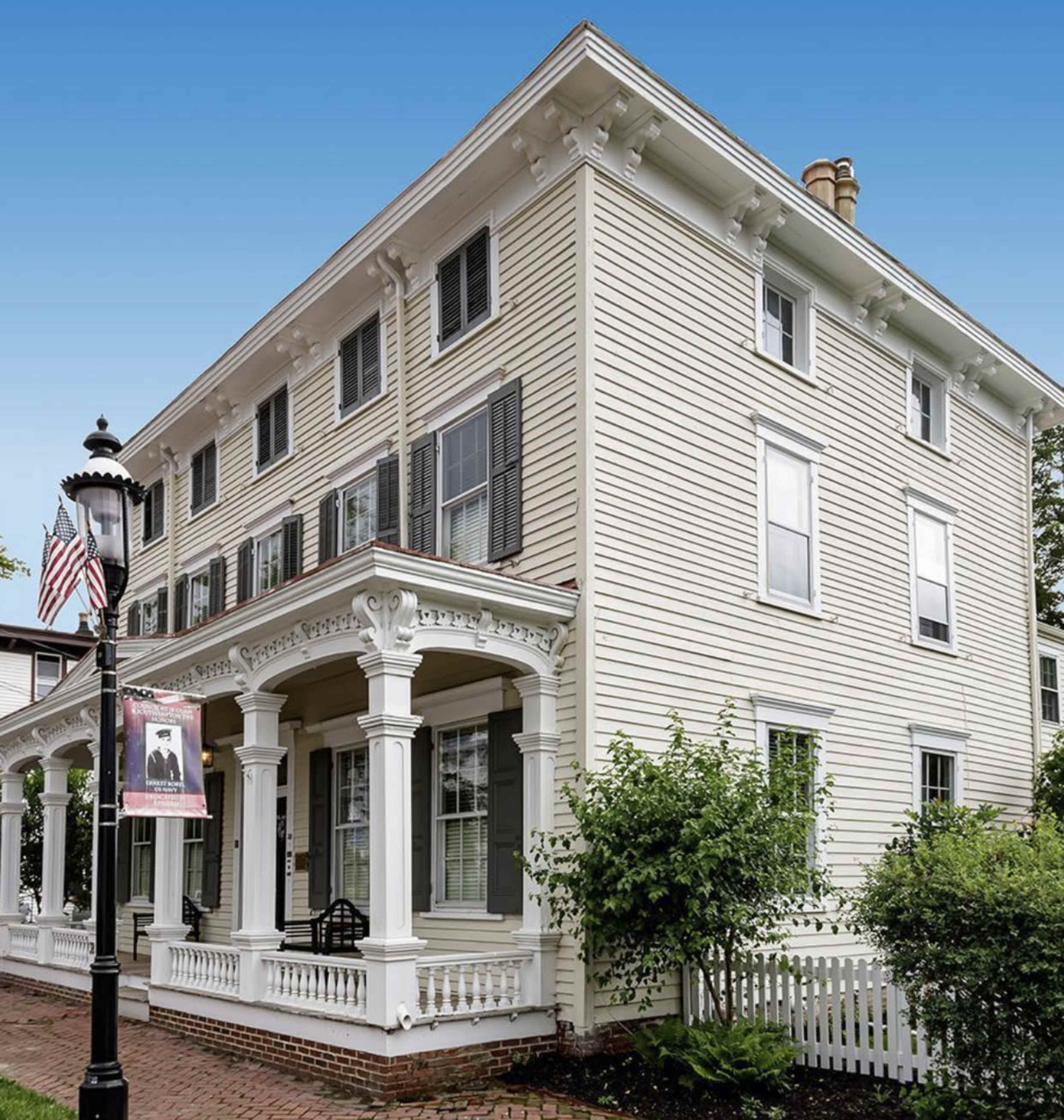 A three-story white house with black shutters features a large porch supported by white columns and is located on a landscaped property.