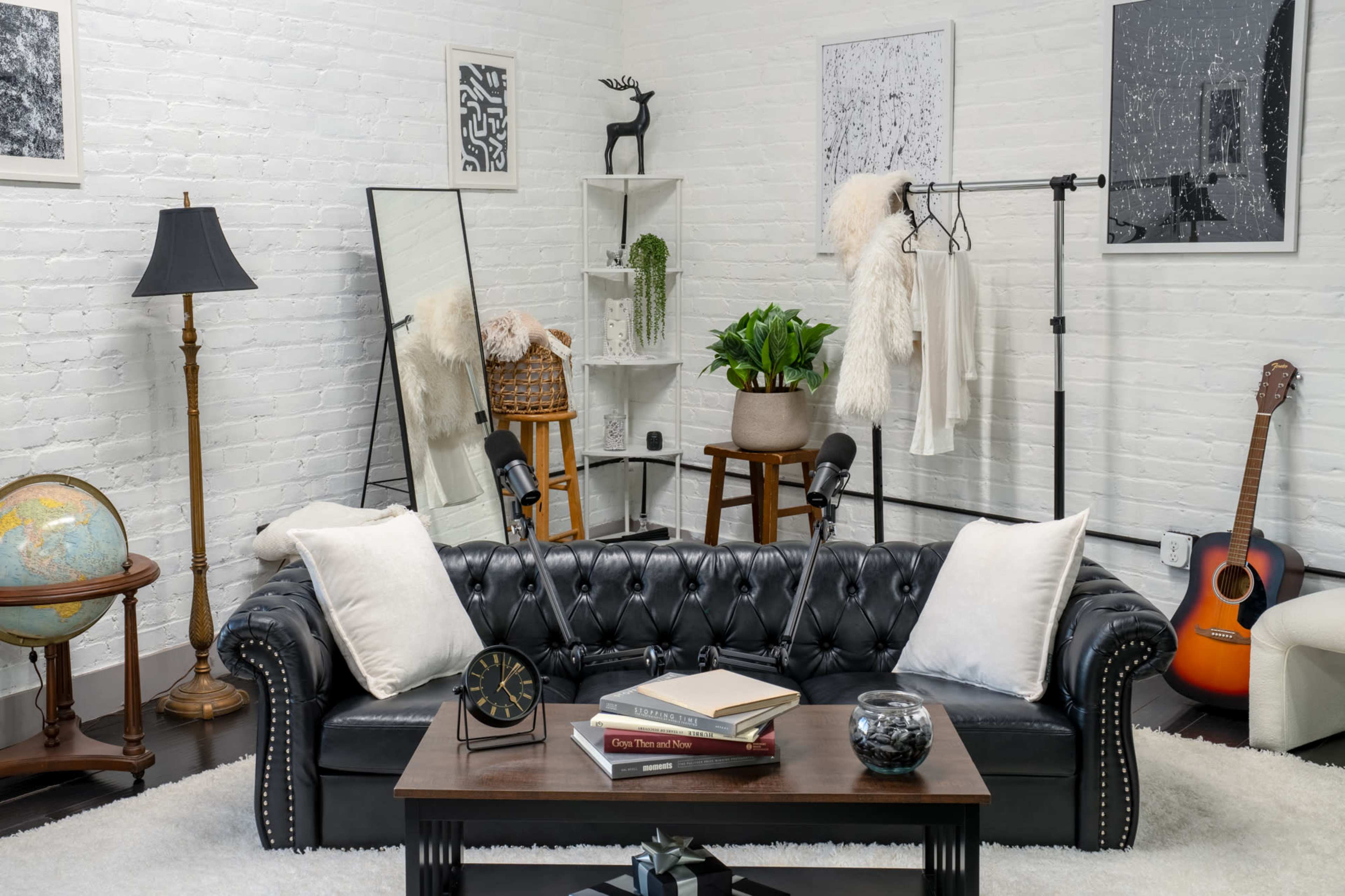 The image shows a modern living space featuring a black leather chesterfield sofa with white pillows, a wooden coffee table, a globe on a stand, and decor items like plants and wall art in a light-colored room.