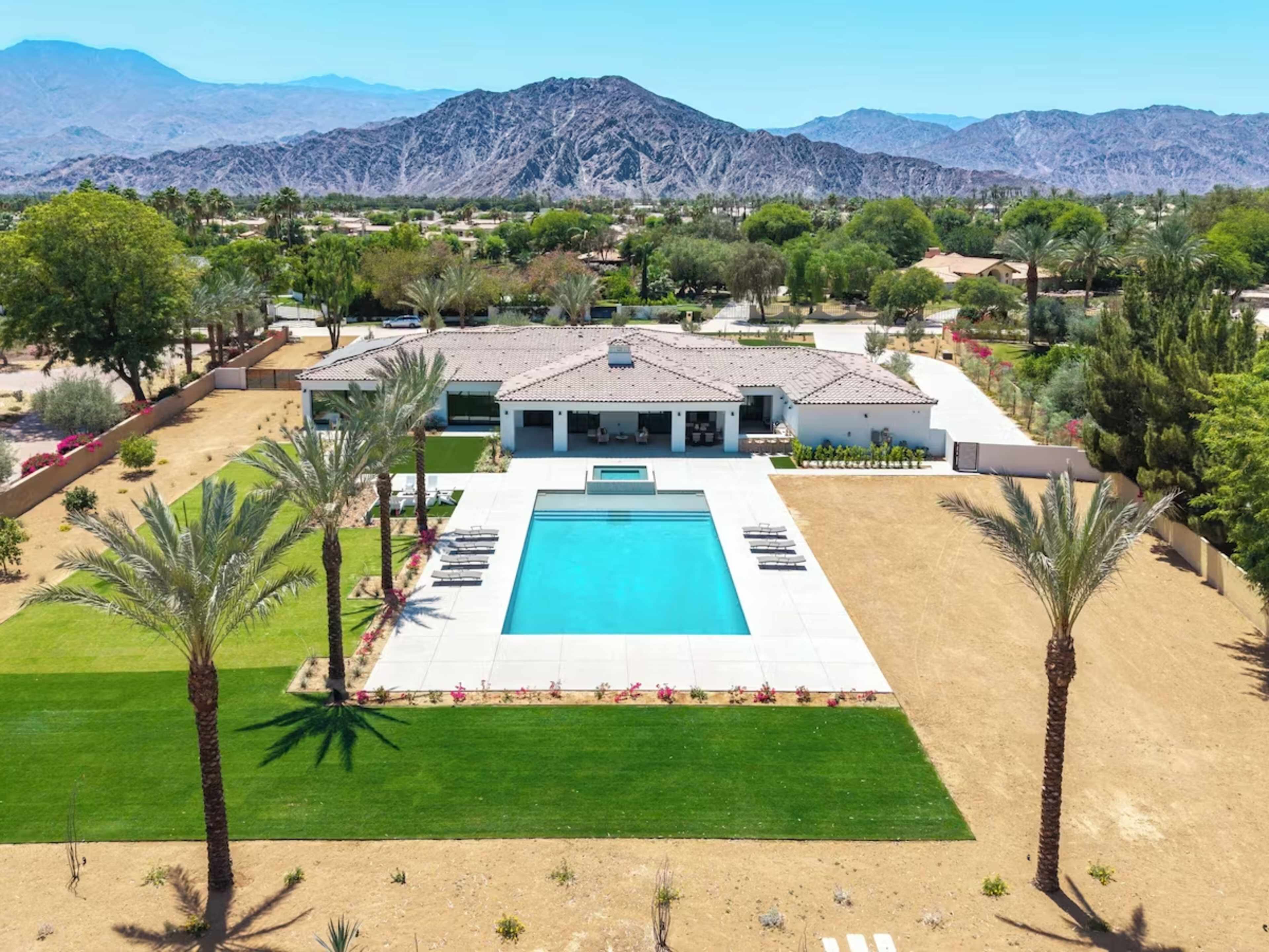 The image shows a modern house with a large pool surrounded by palm trees and landscaped grass, set against a backdrop of mountains.