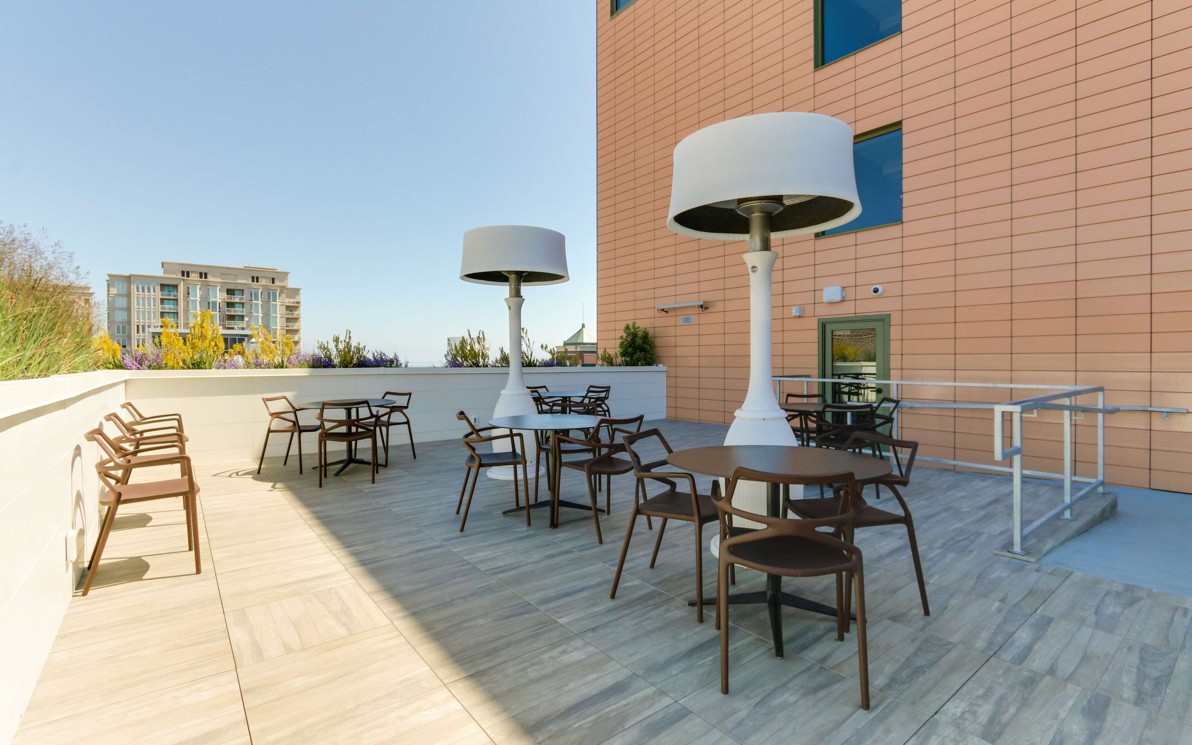 An outdoor patio area with modern tables and chairs, featuring large white lamps, surrounded by a clear sky and adjacent buildings.