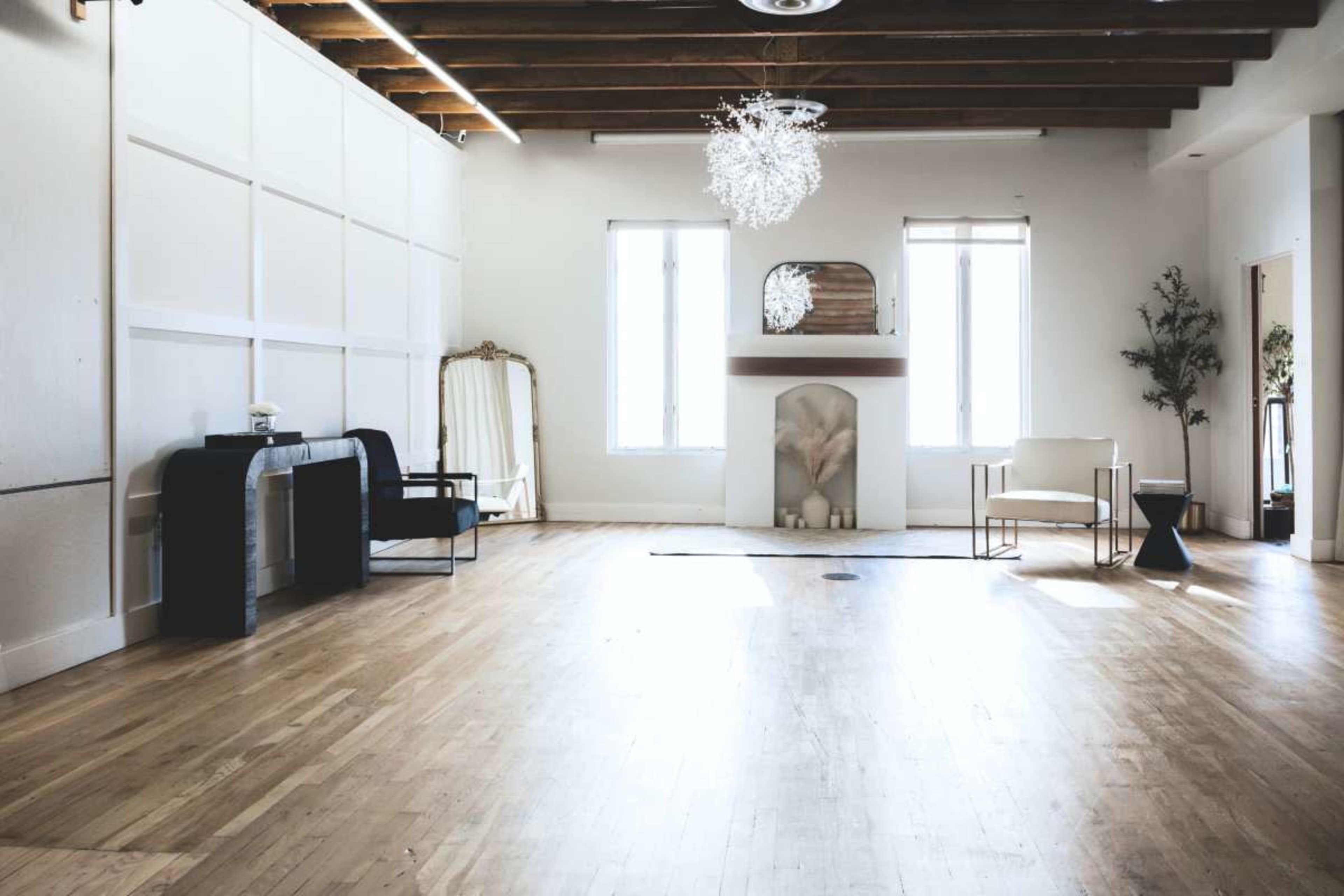The image shows a spacious, well-lit room with wooden floors, large windows, and minimalist furniture, featuring a fireplace and decorative elements.
