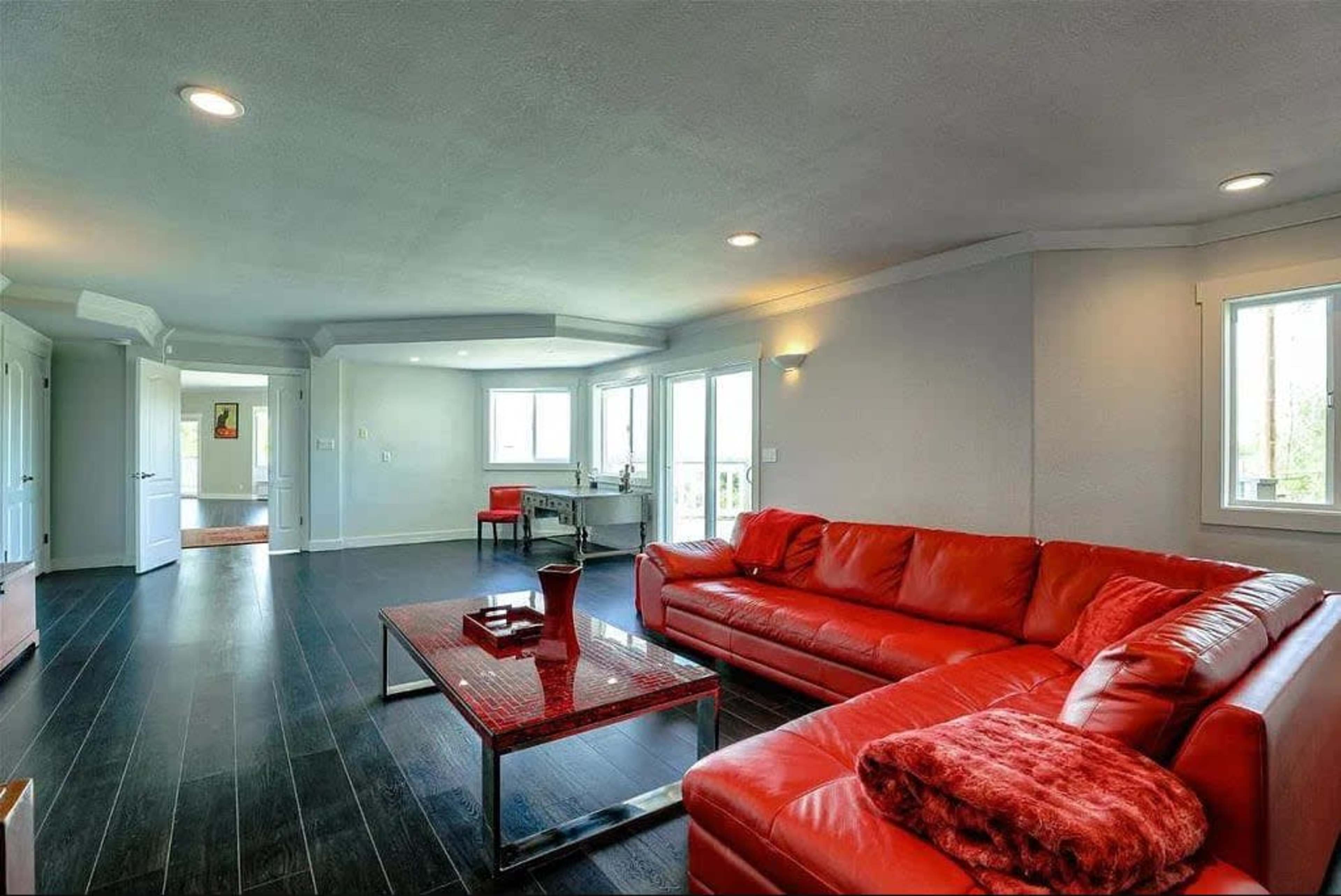 The image shows a spacious living room featuring a red sectional sofa, a glass coffee table, and multiple windows allowing natural light to enter.