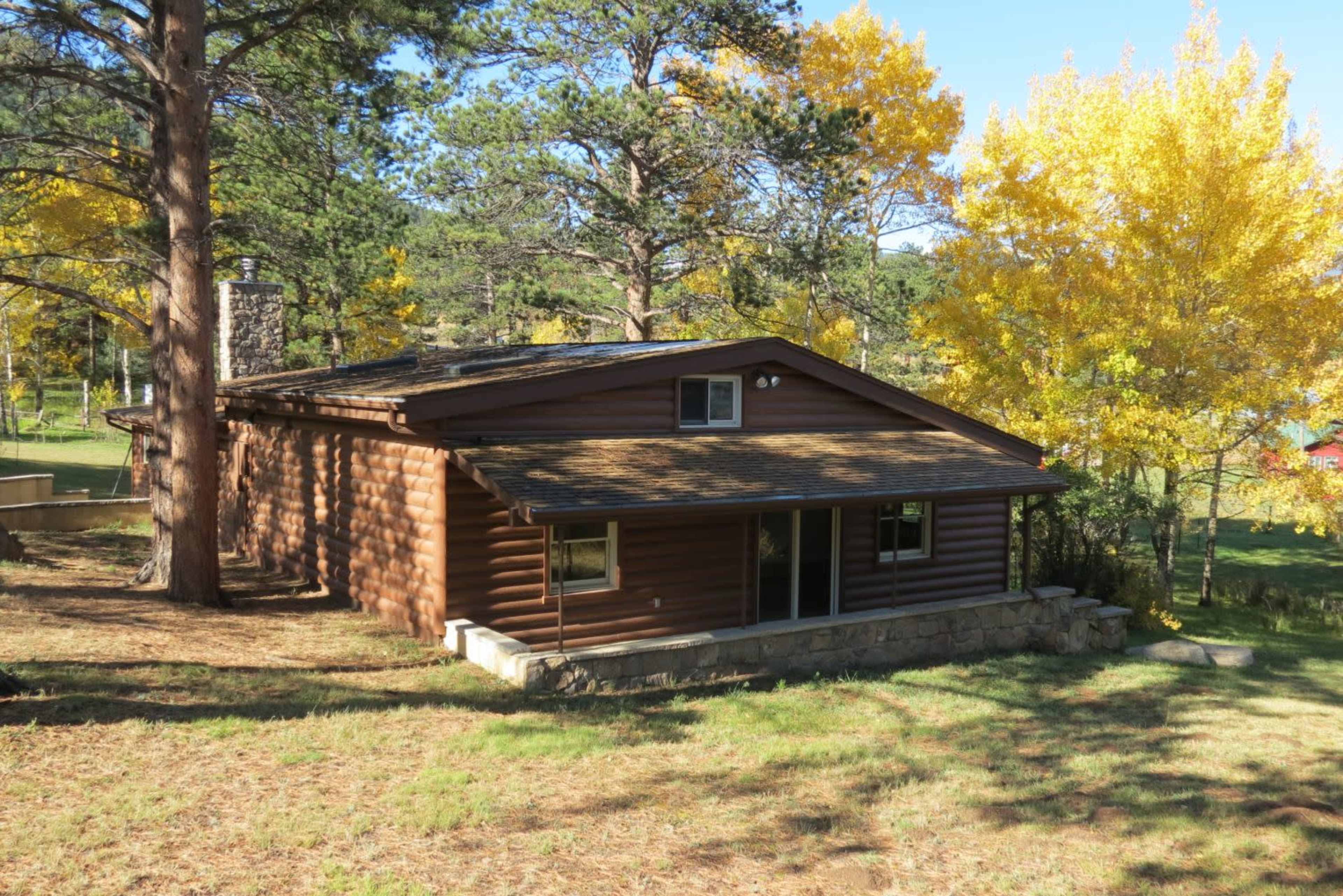 A wooden cabin is situated on a grassy slope surrounded by trees with autumn foliage.