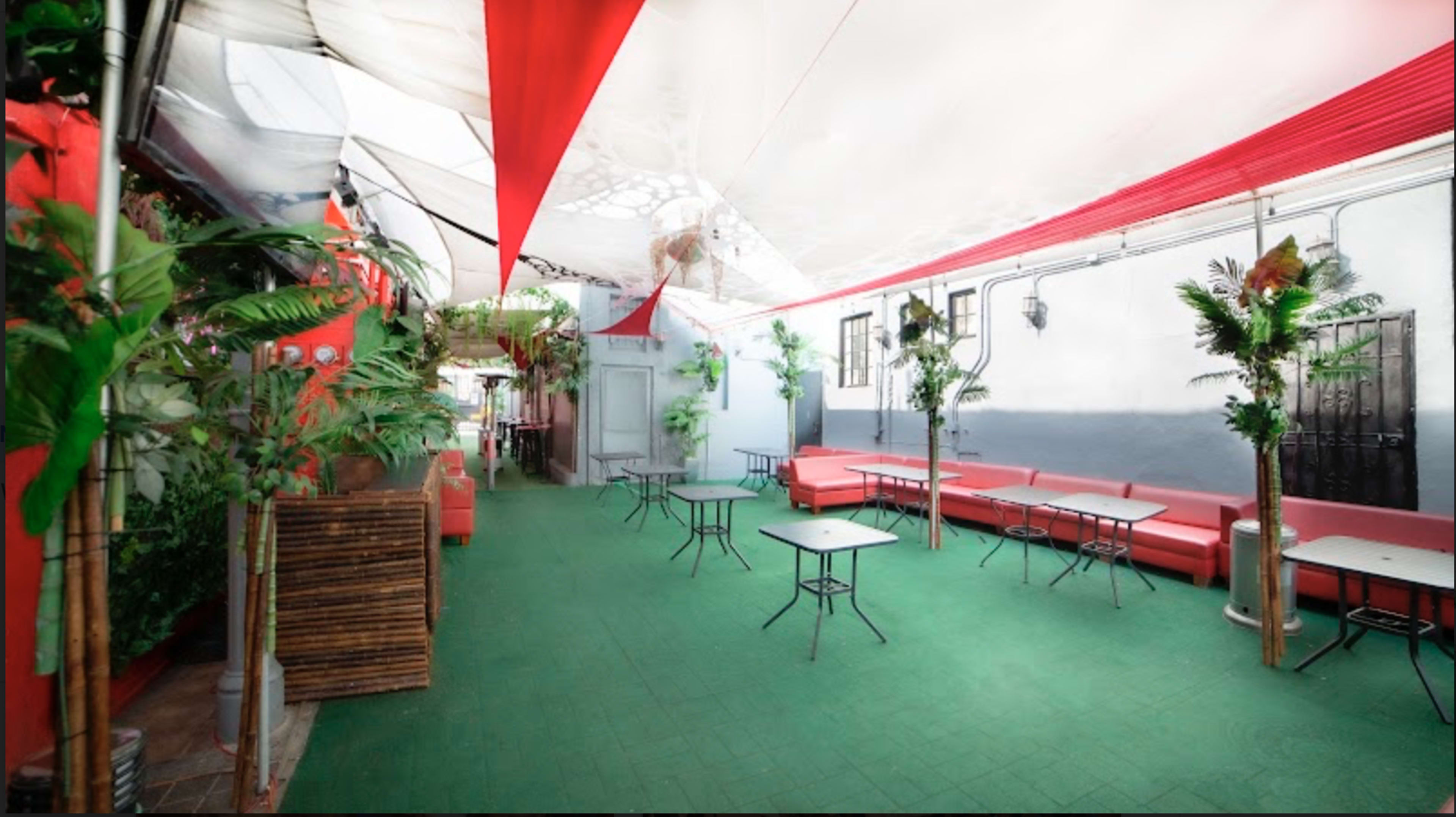 A covered outdoor area with red decor, green flooring, and small tables surrounded by potted plants.