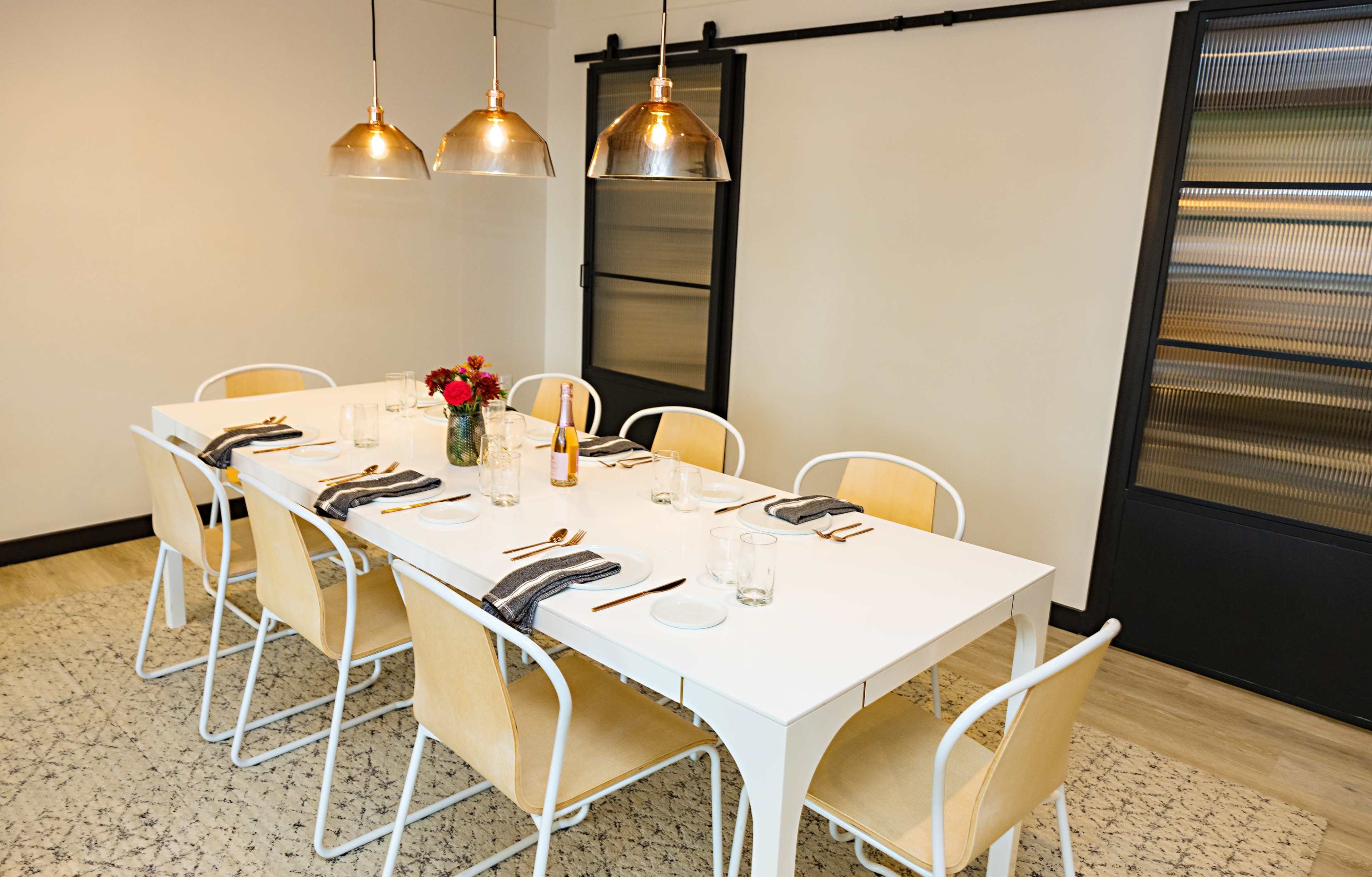 A long white dining table is set with utensils and glasses in a modern room featuring pendant lights and a glass door.