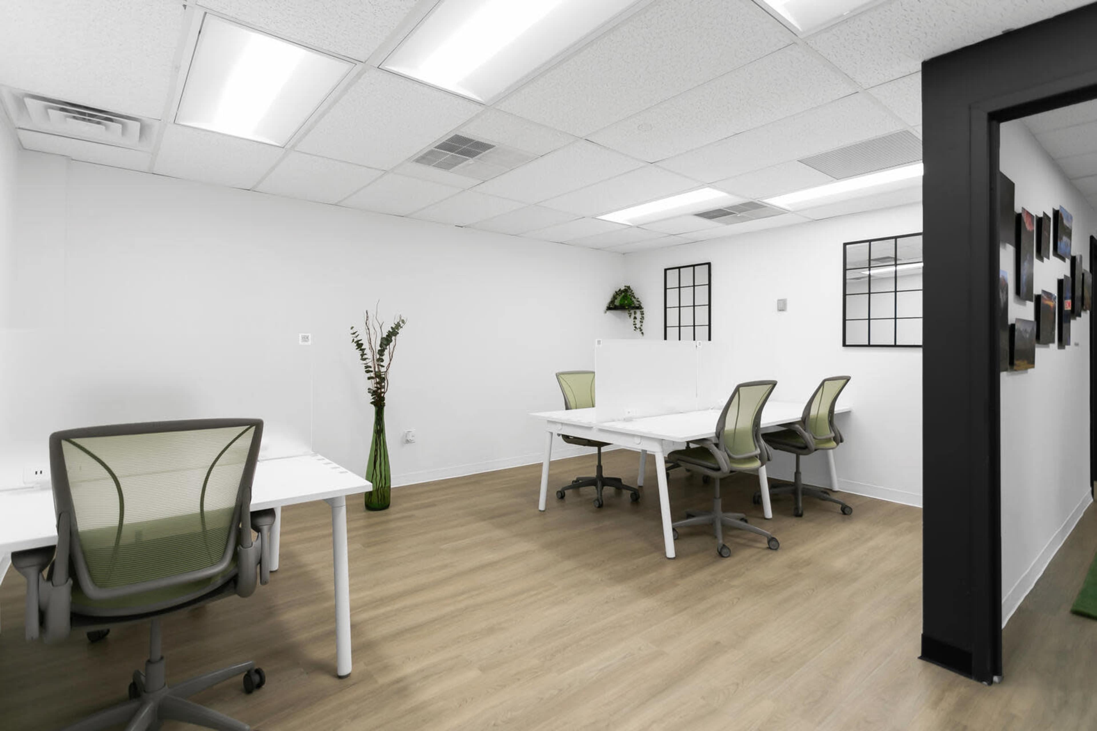 The image depicts a modern, minimally designed office space with four desks and ergonomic chairs arranged neatly on a light wooden floor.