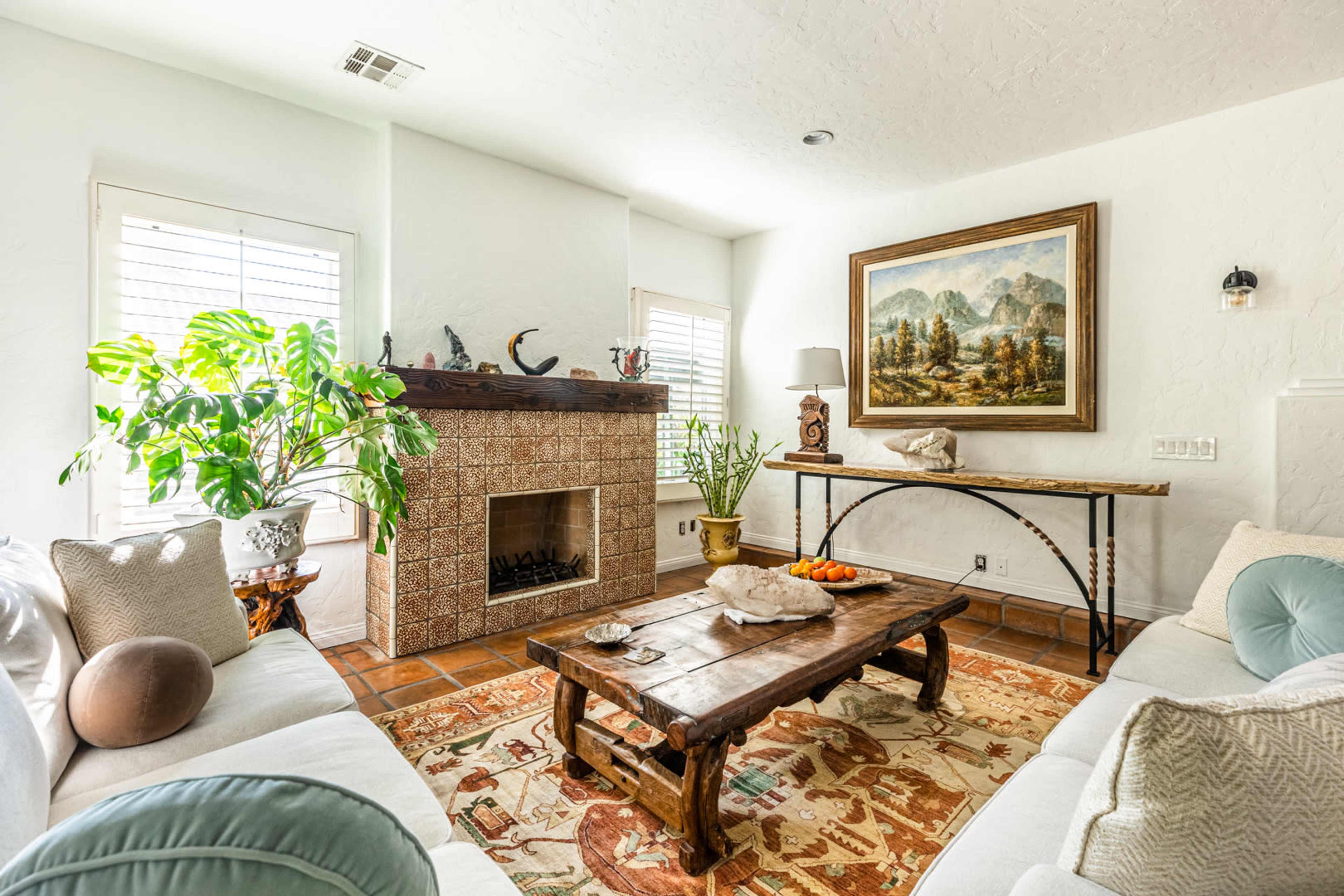 A bright living room features a fireplace, a large painting of mountains, a wooden coffee table with fruit, and a green plant beside a sofa.