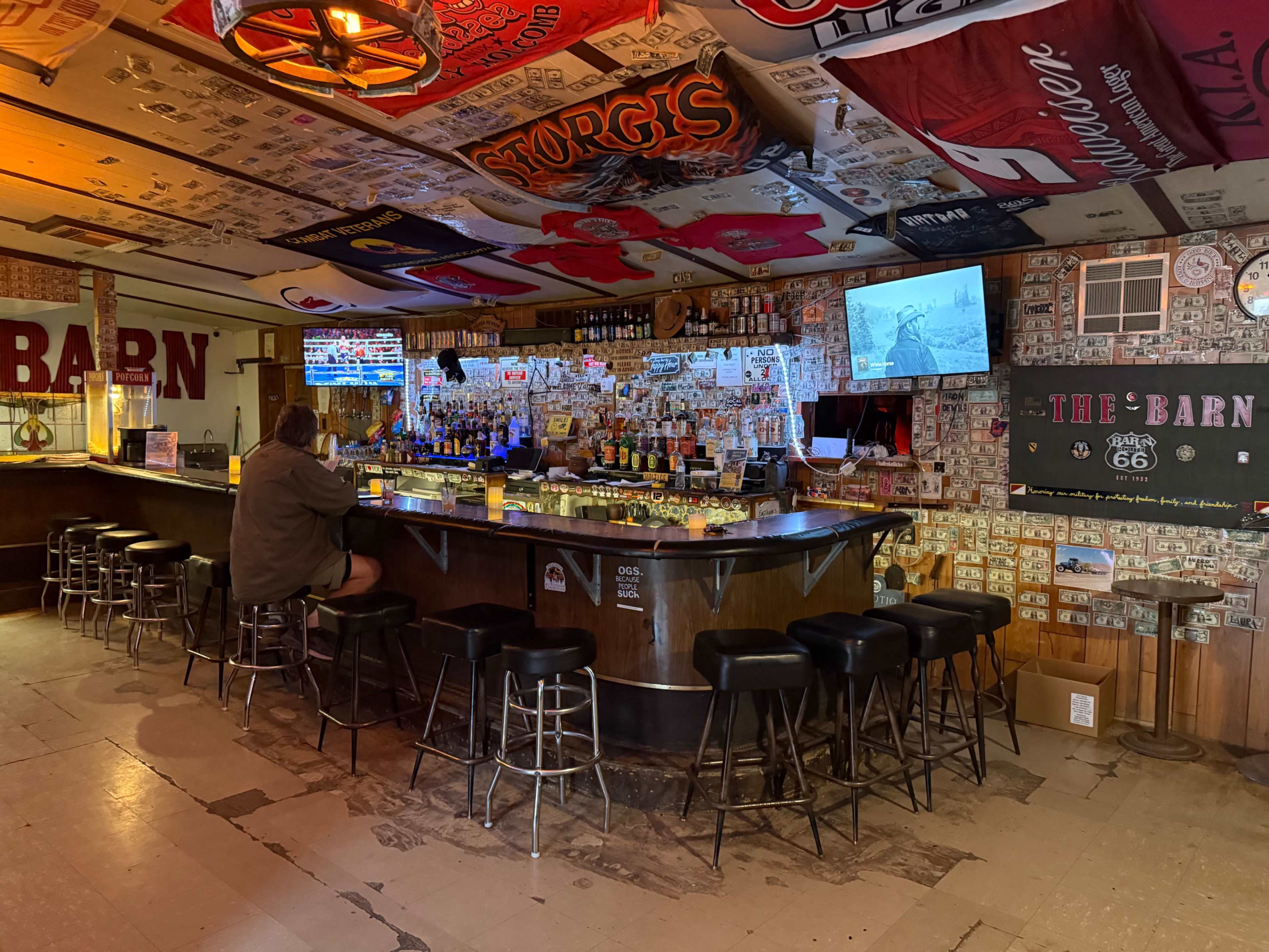 A person sits at the bar of a rustic establishment adorned with various stickers and memorabilia on the walls and ceiling.