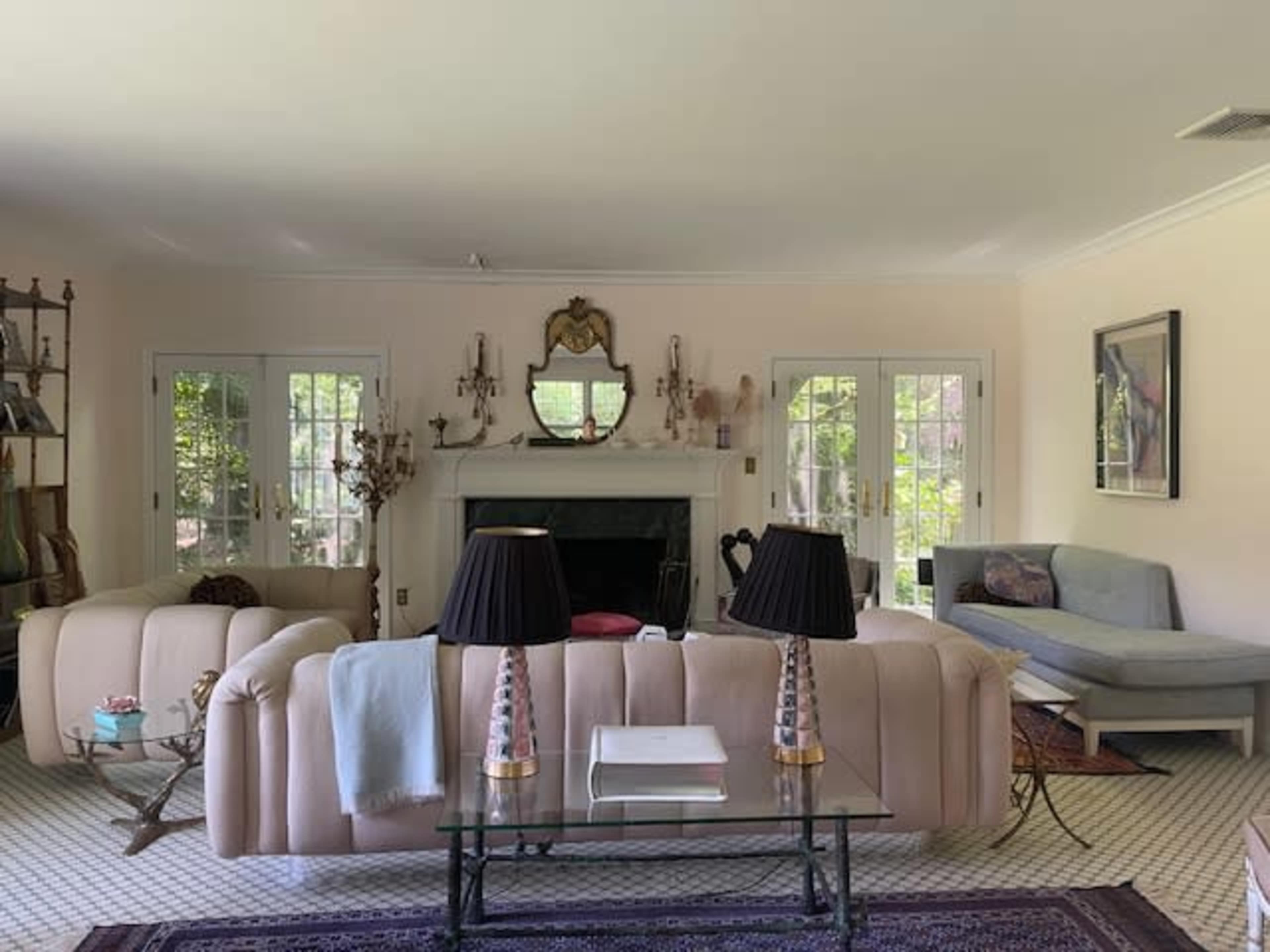 The living room features two pale sofas facing a glass coffee table, with a fireplace and large windows allowing natural light.