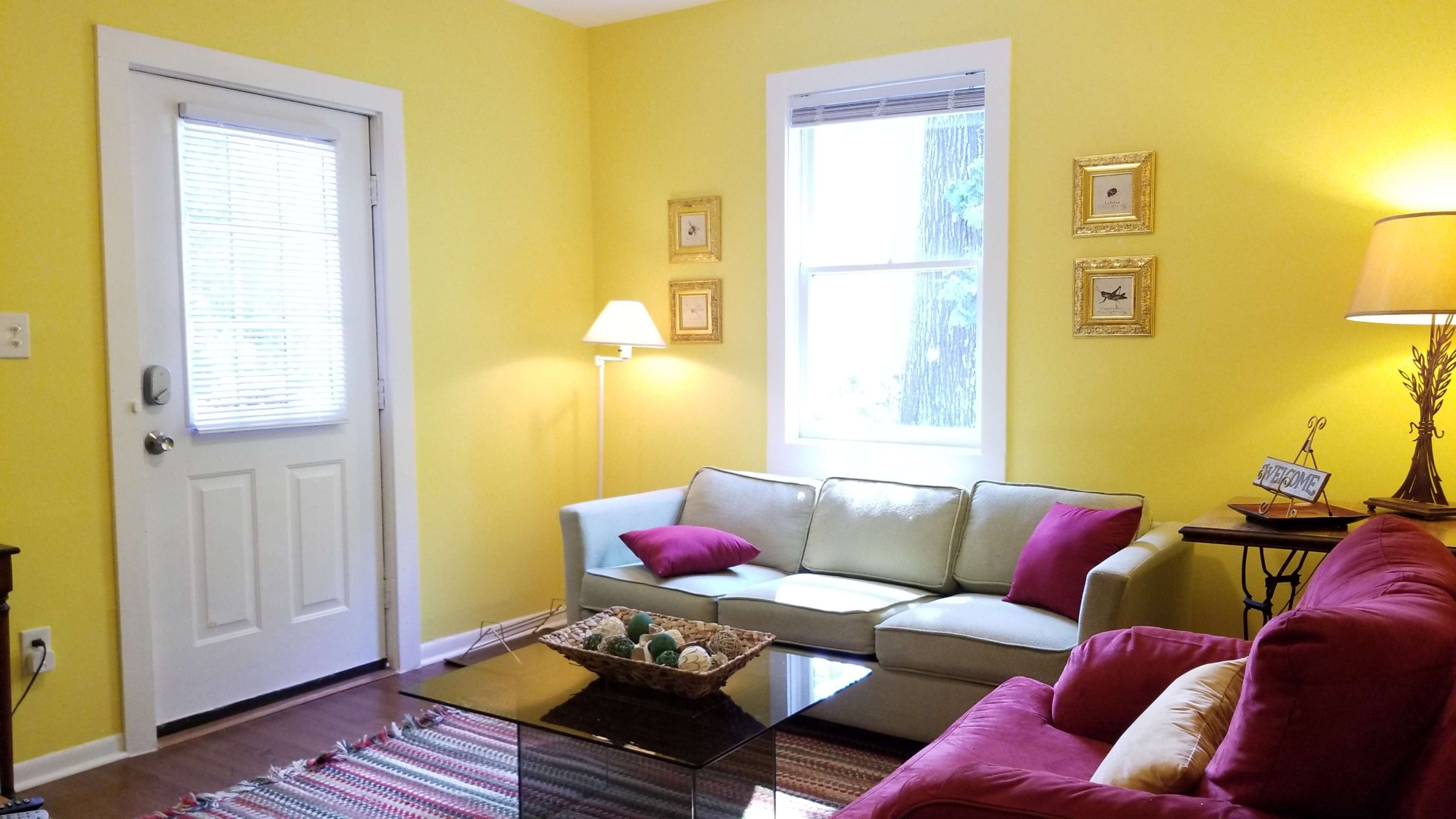 A living room with yellow walls, a light-colored sofa adorned with pink pillows, a red couch, a glass coffee table, a floor lamp, and framed pictures on the walls.