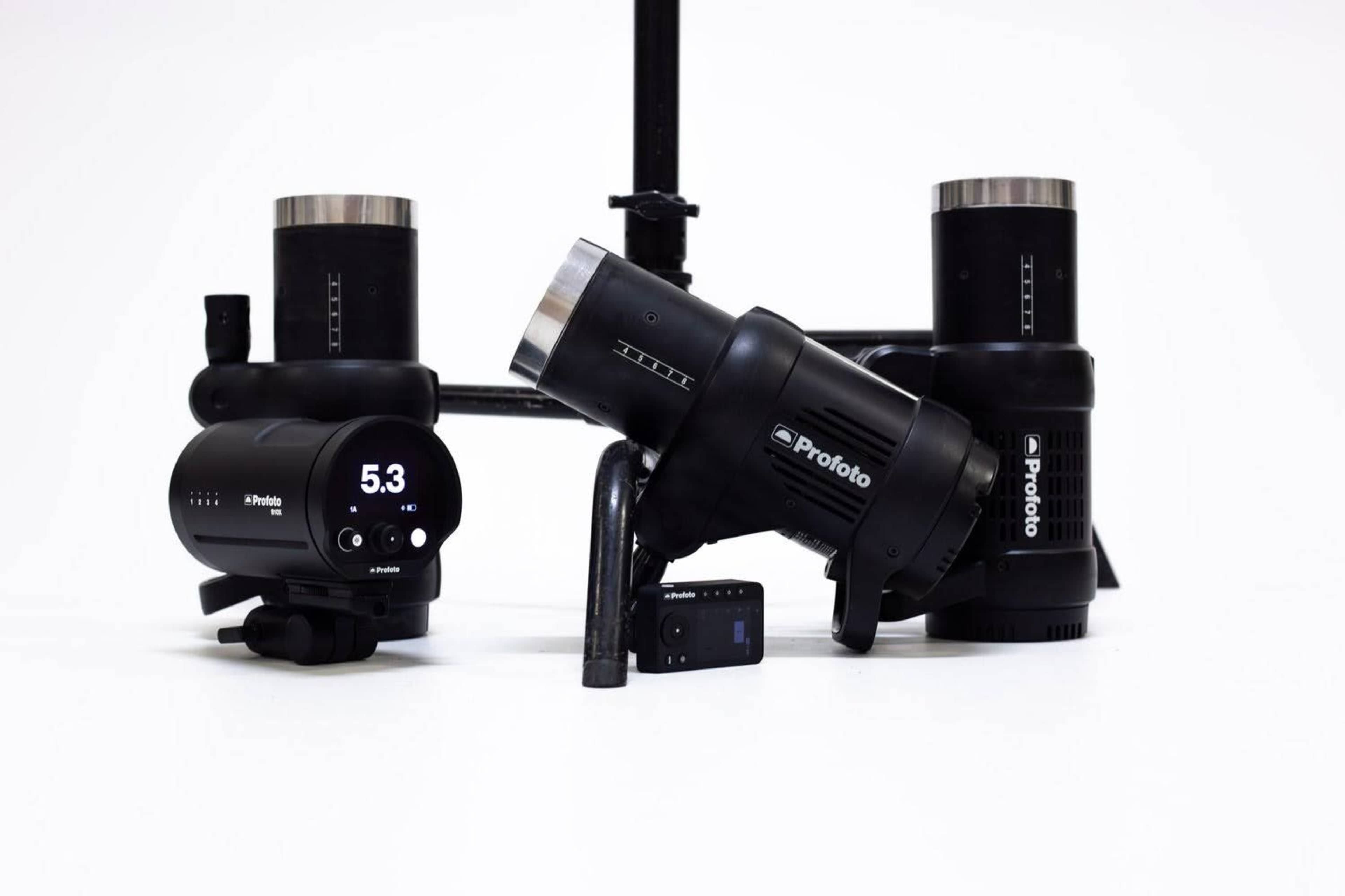 The image shows a collection of Profoto lighting equipment arranged on a plain white background, including a flash head, battery pack, and several modifiers.