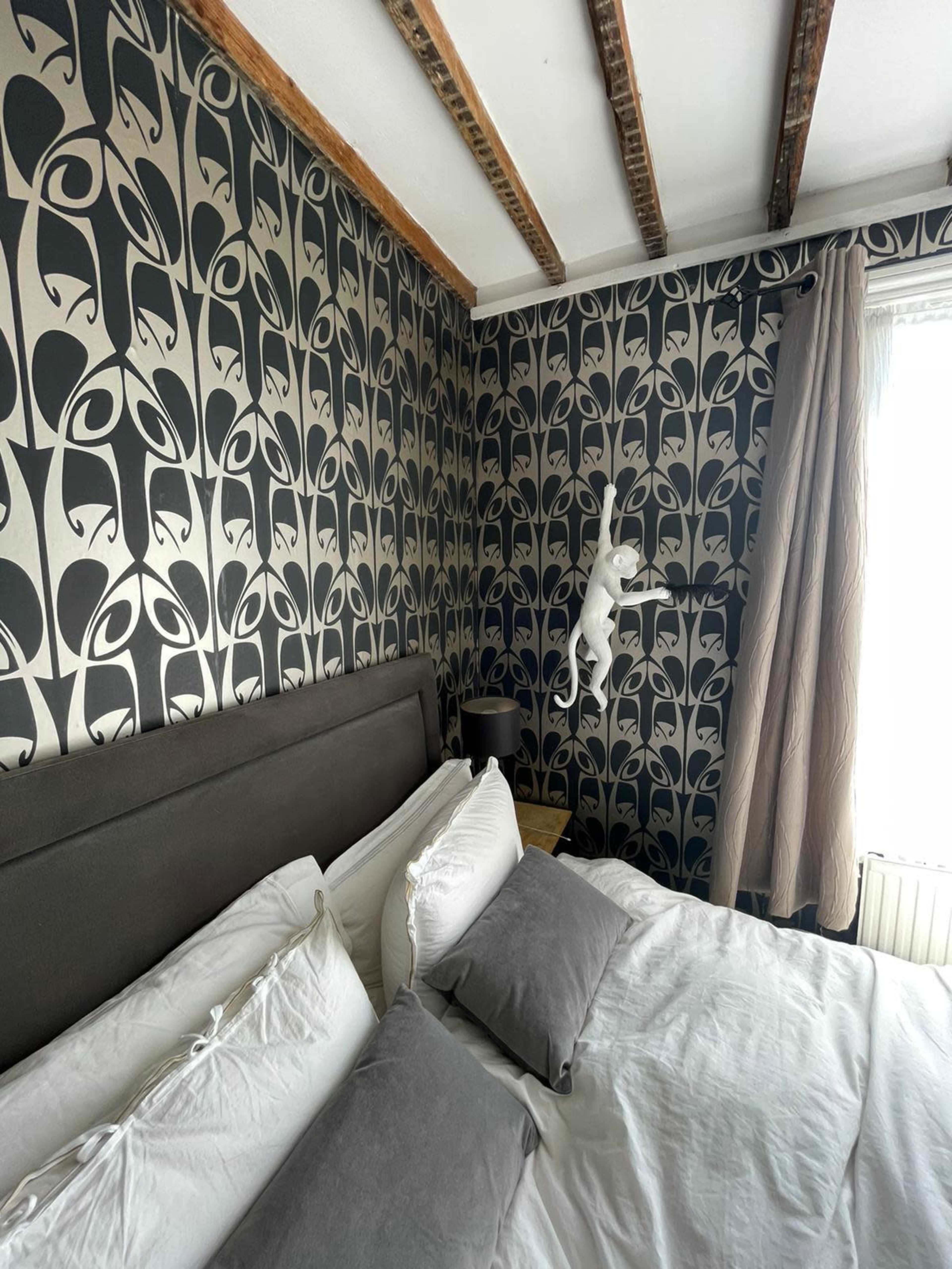 A cat climbs a patterned wall in a bedroom with a large bed and wooden ceiling beams.