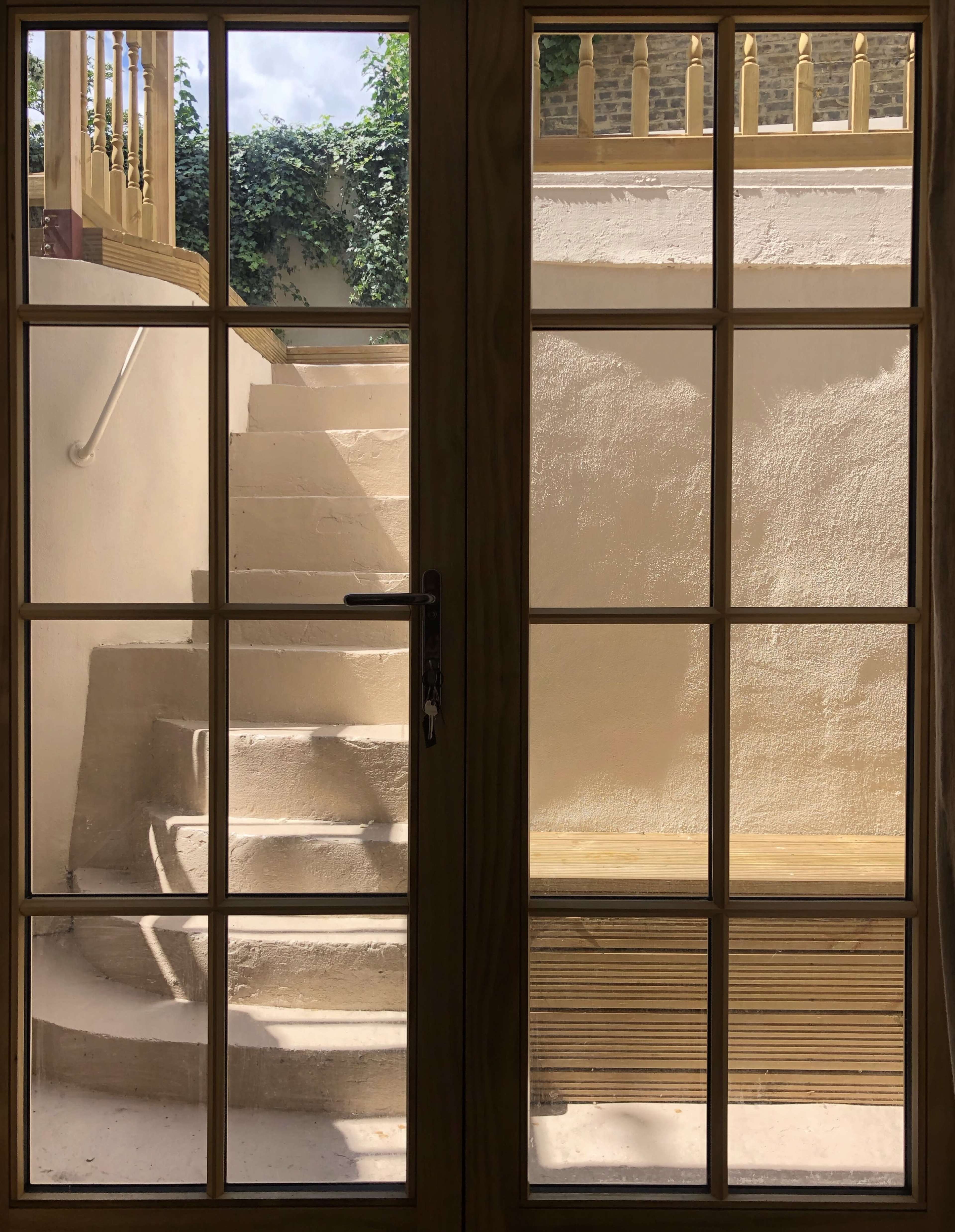 A view through a wooden-framed glass door shows a staircase leading up to a deck surrounded by greenery.