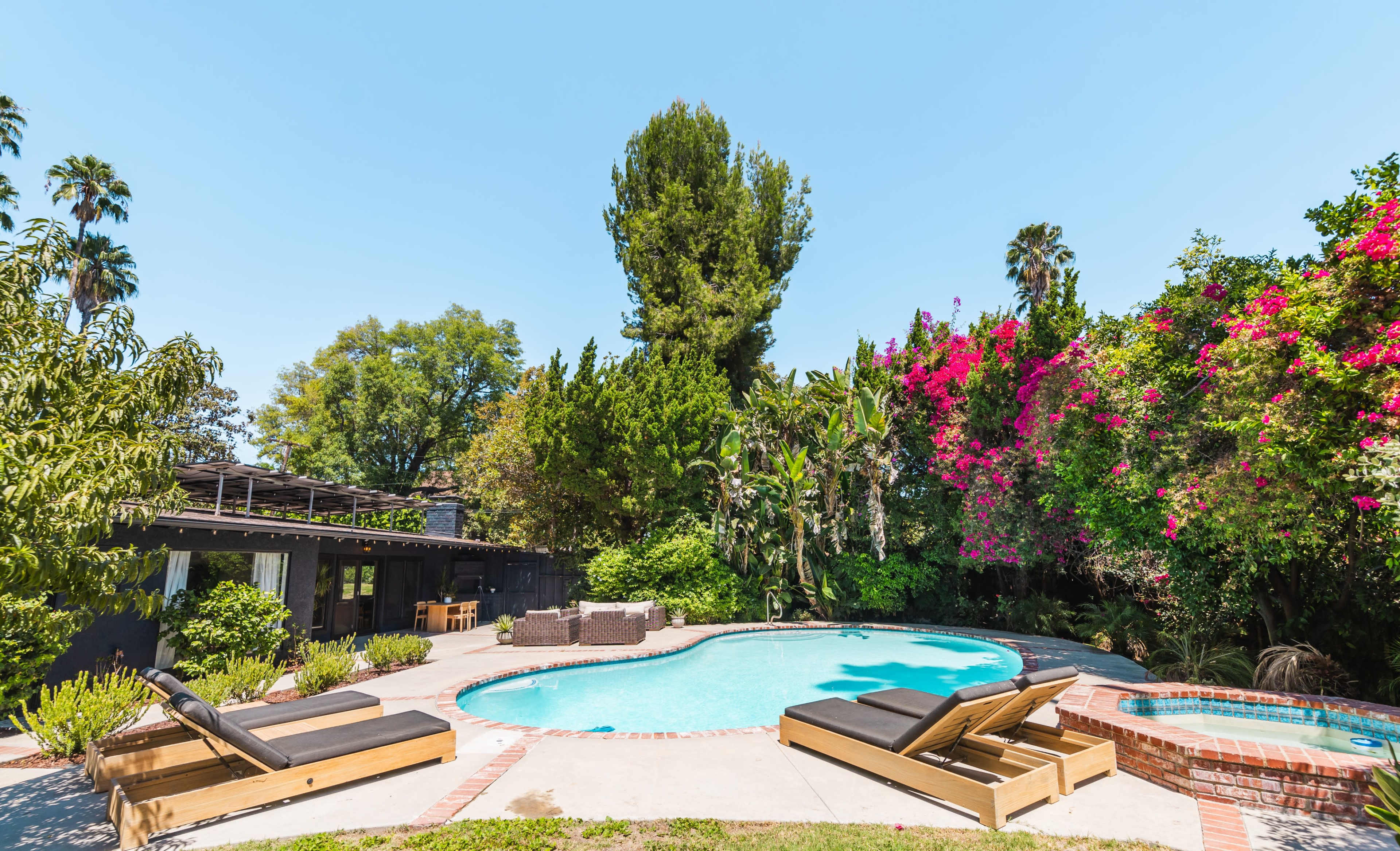 A swimming pool surrounded by lounge chairs and lush tropical foliage, with a backdrop of flowering plants and trees.