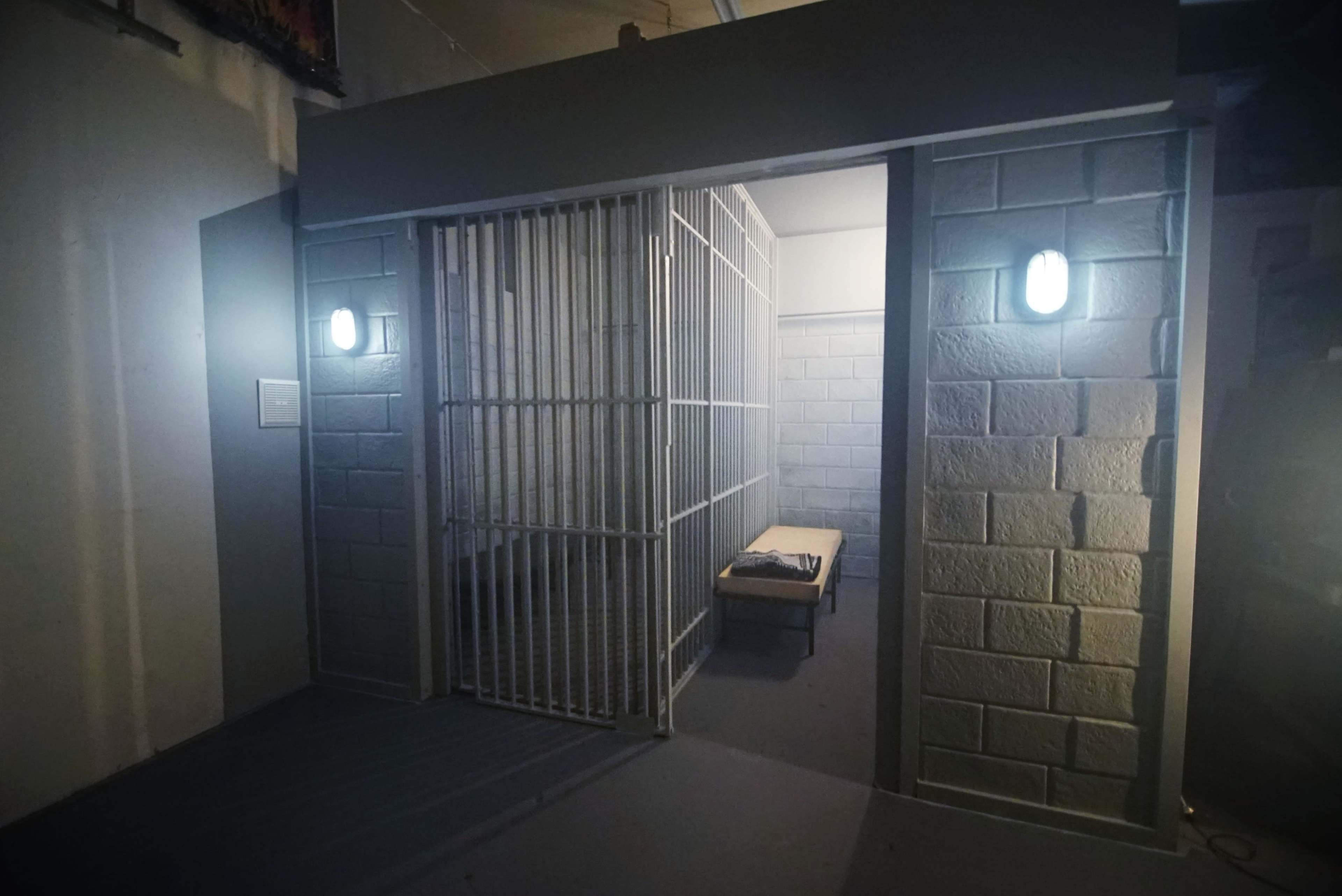 A small jail cell with a barred door and a bench inside, illuminated by two wall-mounted lights.
