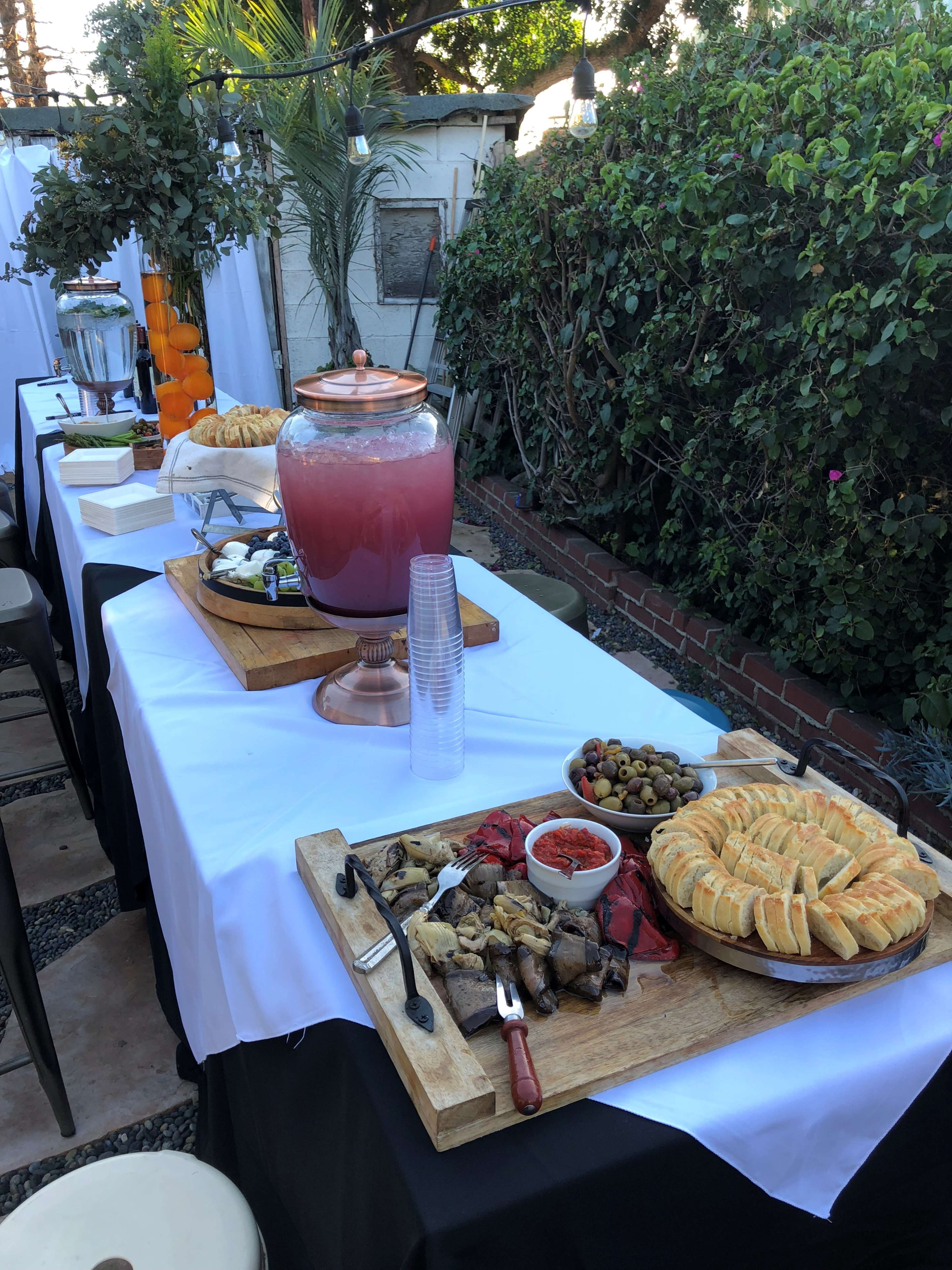 A table is set with various food items, including pastries, olives, and a drink dispenser, surrounded by greenery.