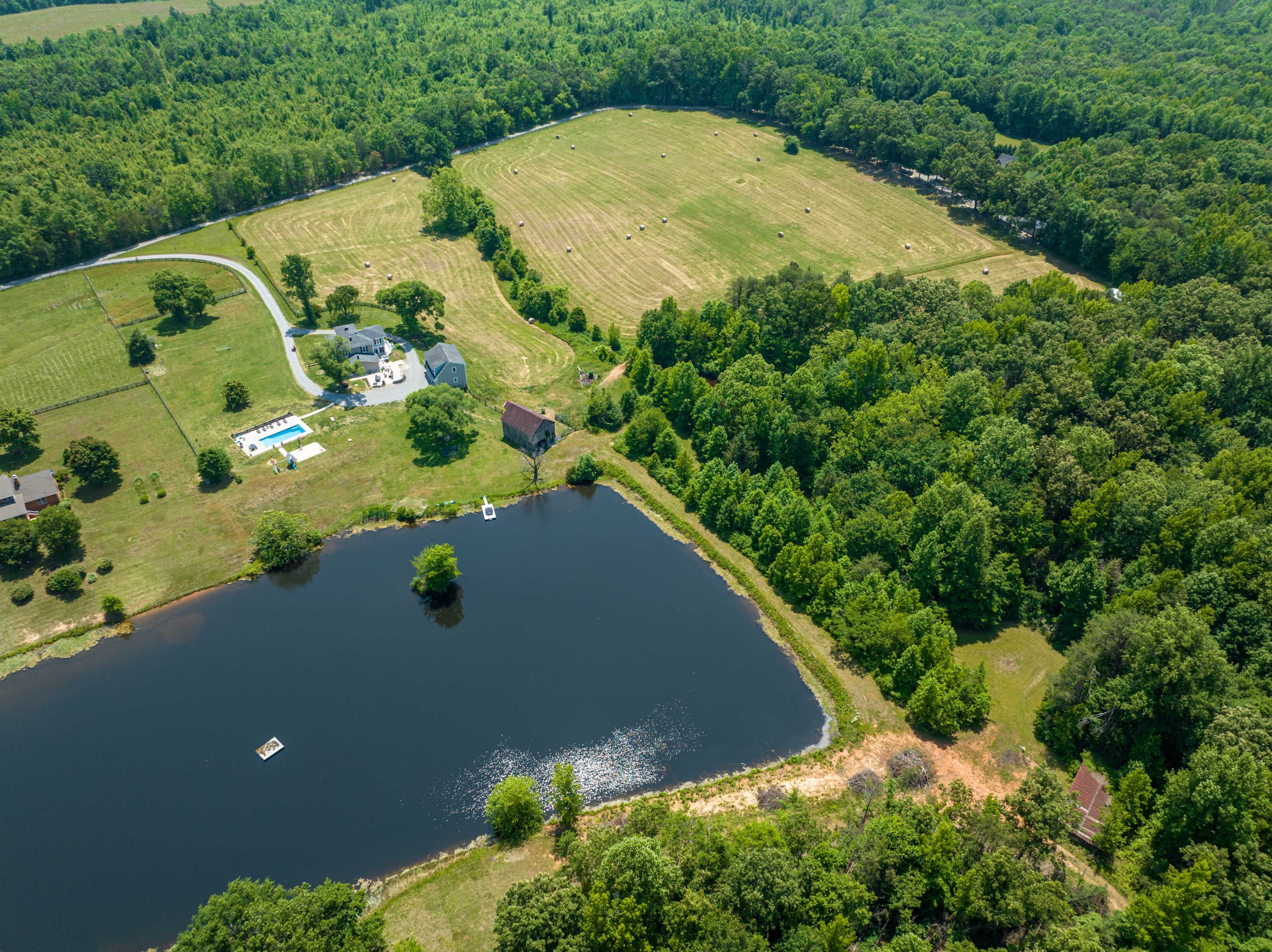 The image shows an aerial view of a rural landscape featuring a pond, a residential area with houses, a swimming pool, and fields with hay bales, surrounded by greenery.