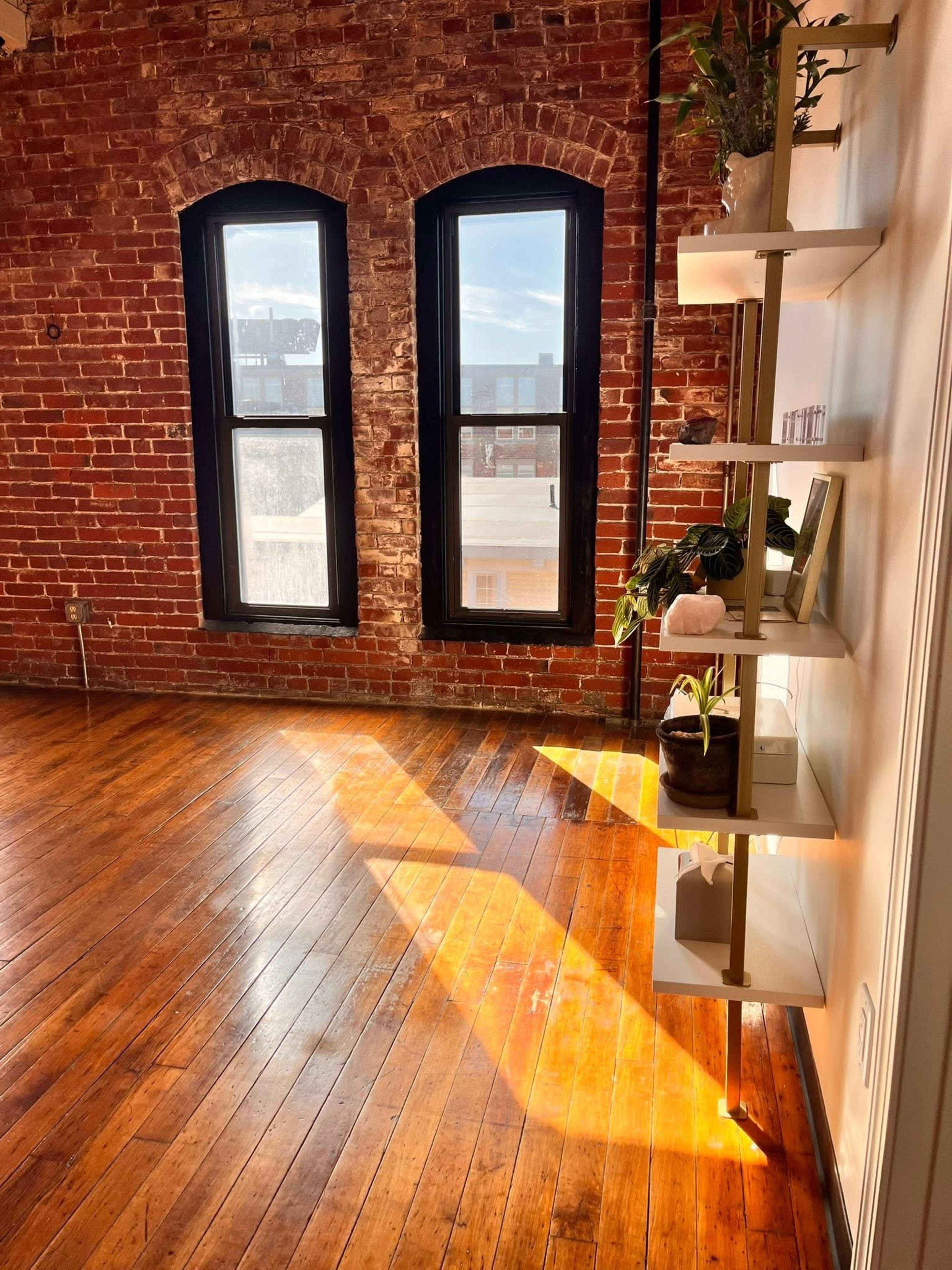 A sunlit room features exposed brick walls, large windows, and a shelf with plants against a wooden floor.