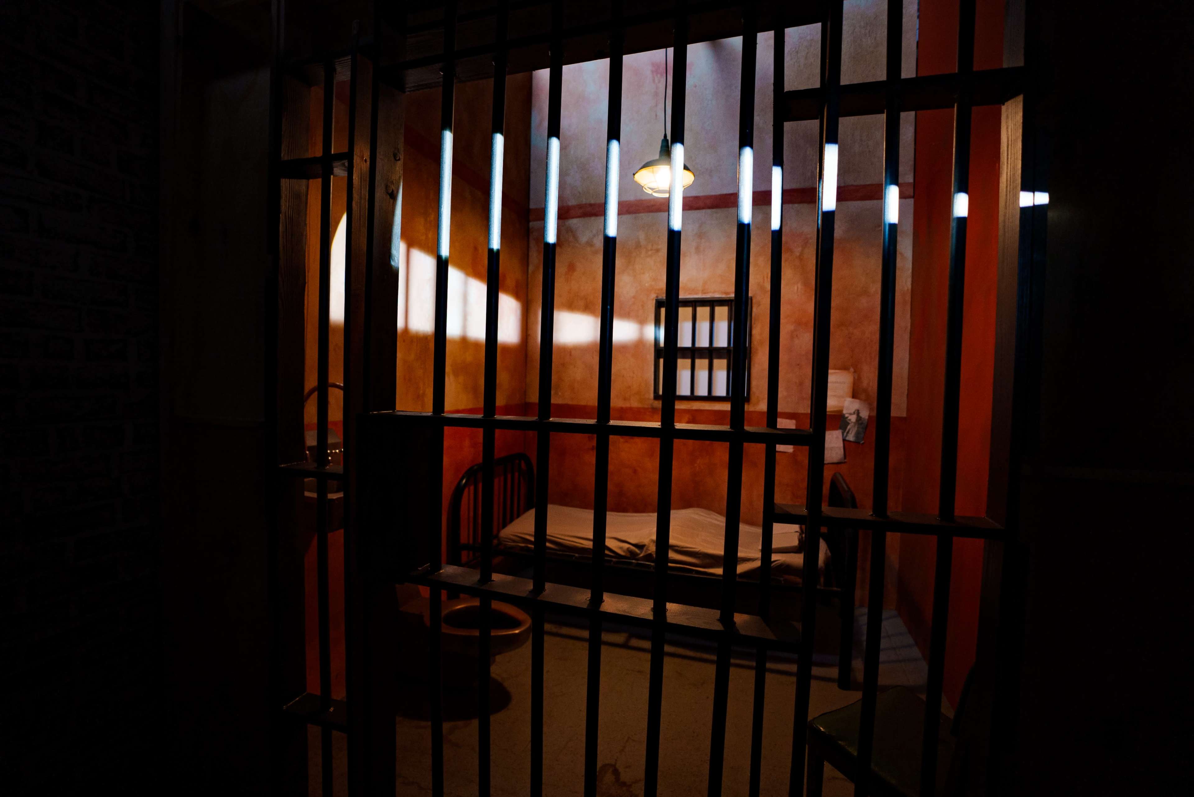 A jail cell with bars, a bed, and a small window.