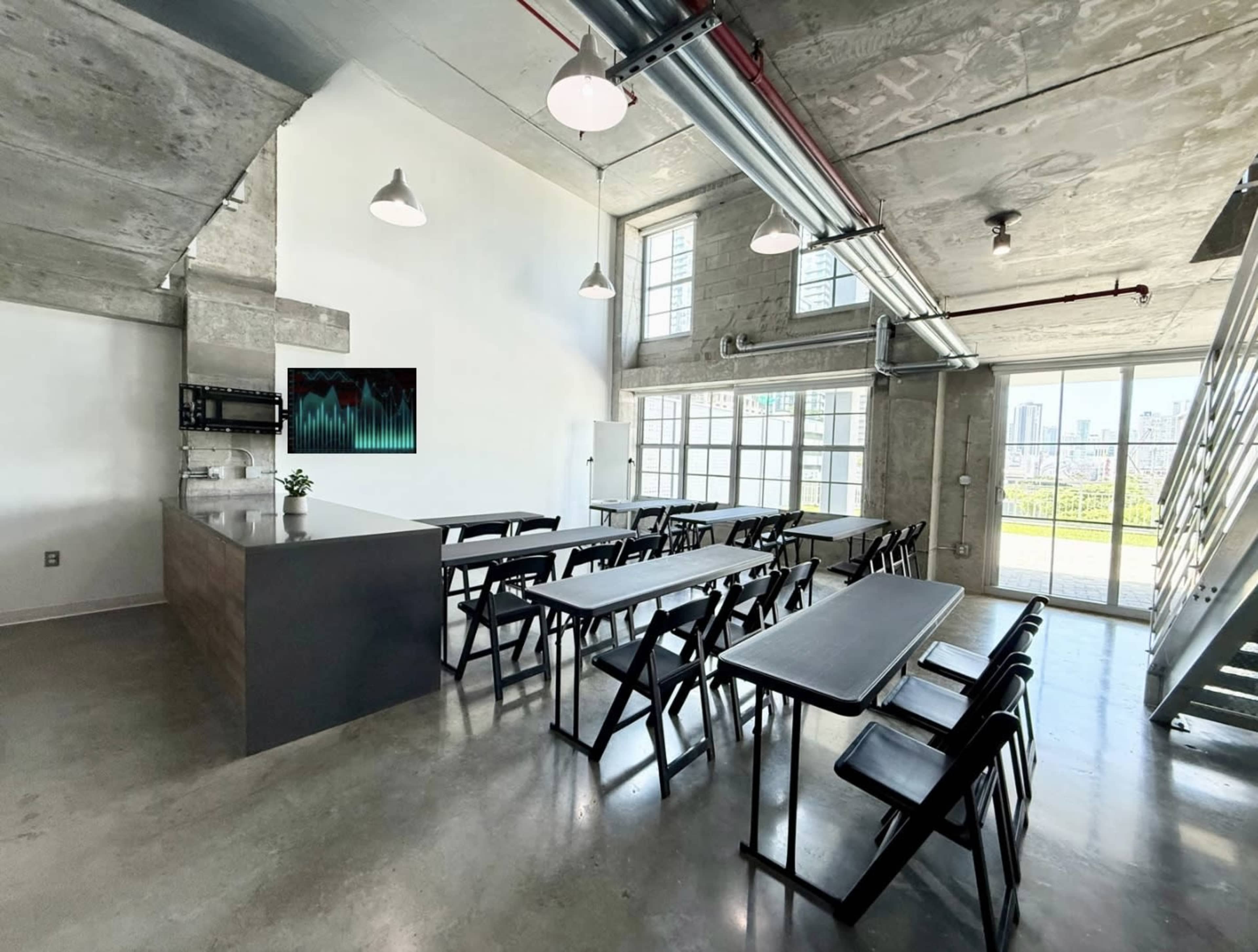 The image shows a modern industrial-style room with concrete walls, long rectangular tables, and large windows offering a view of the city.