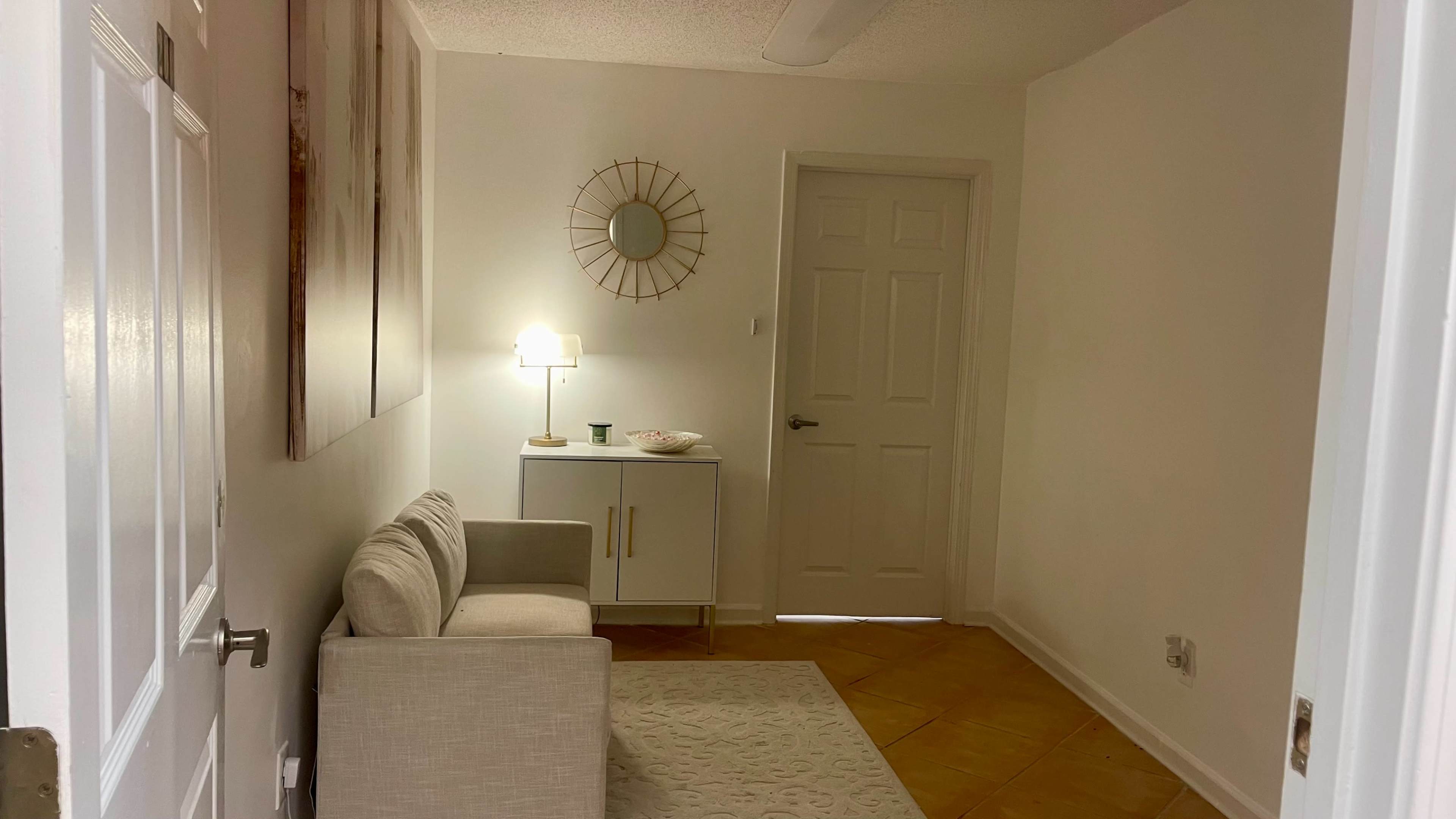A small, brightly lit room features a light-colored couch, a side table with a lamp, and a wall-mounted mirror.