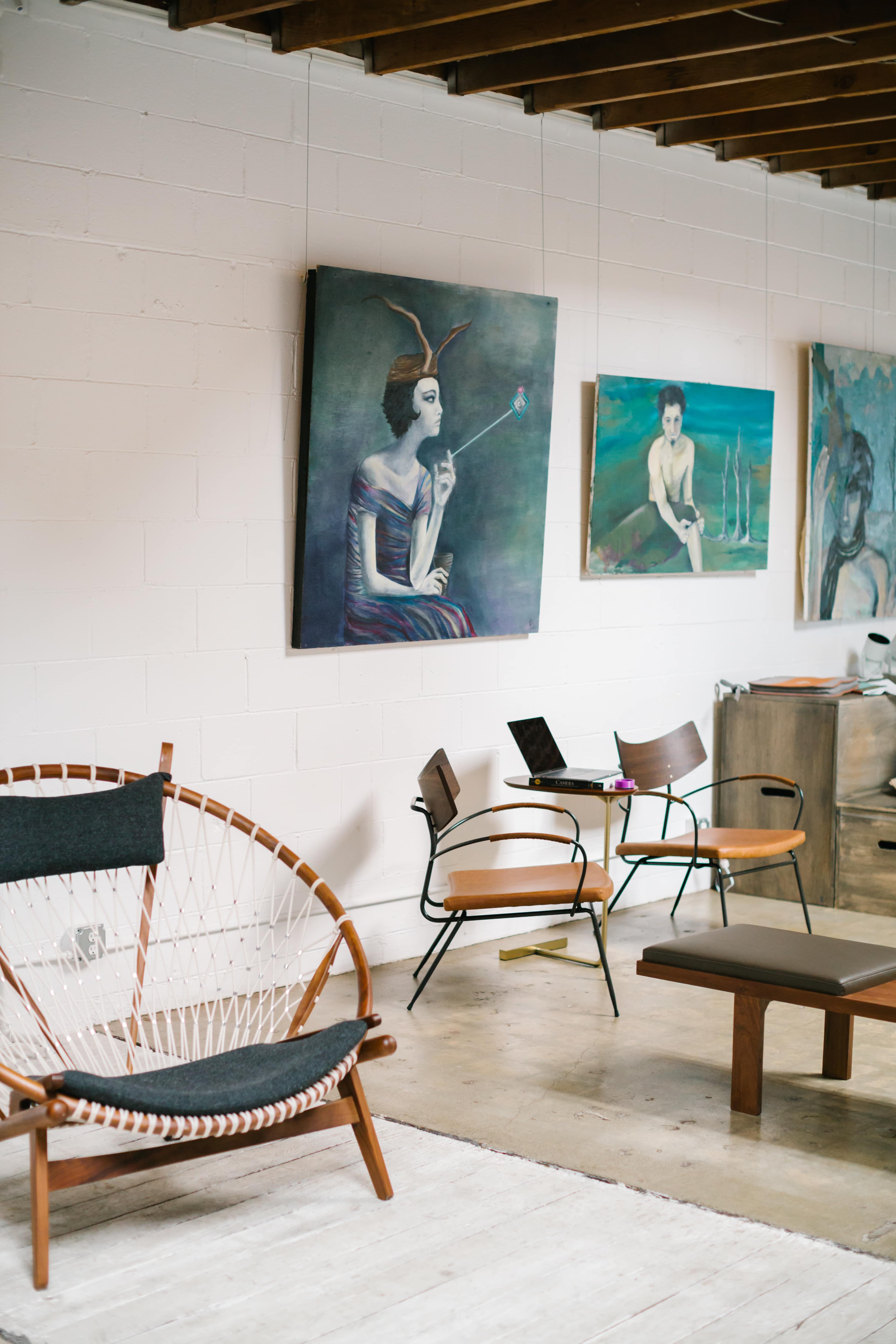 The image shows an art gallery interior with several large paintings on the white walls and modern furniture, including a unique chair and wooden bench.