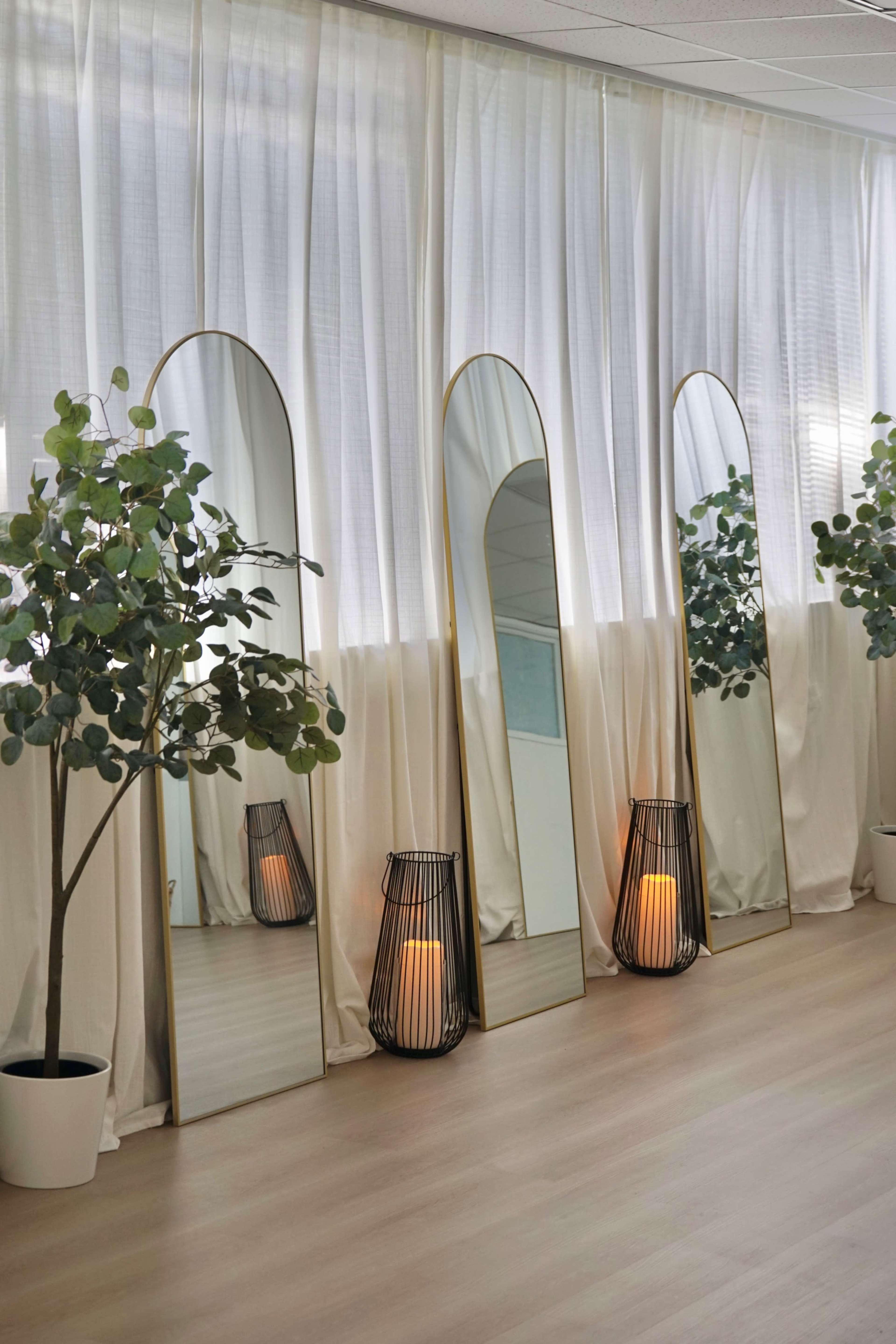 The image features three tall, arched mirrors placed against a backdrop of sheer white curtains, with potted plants and lanterns positioned beside them on a wooden floor.