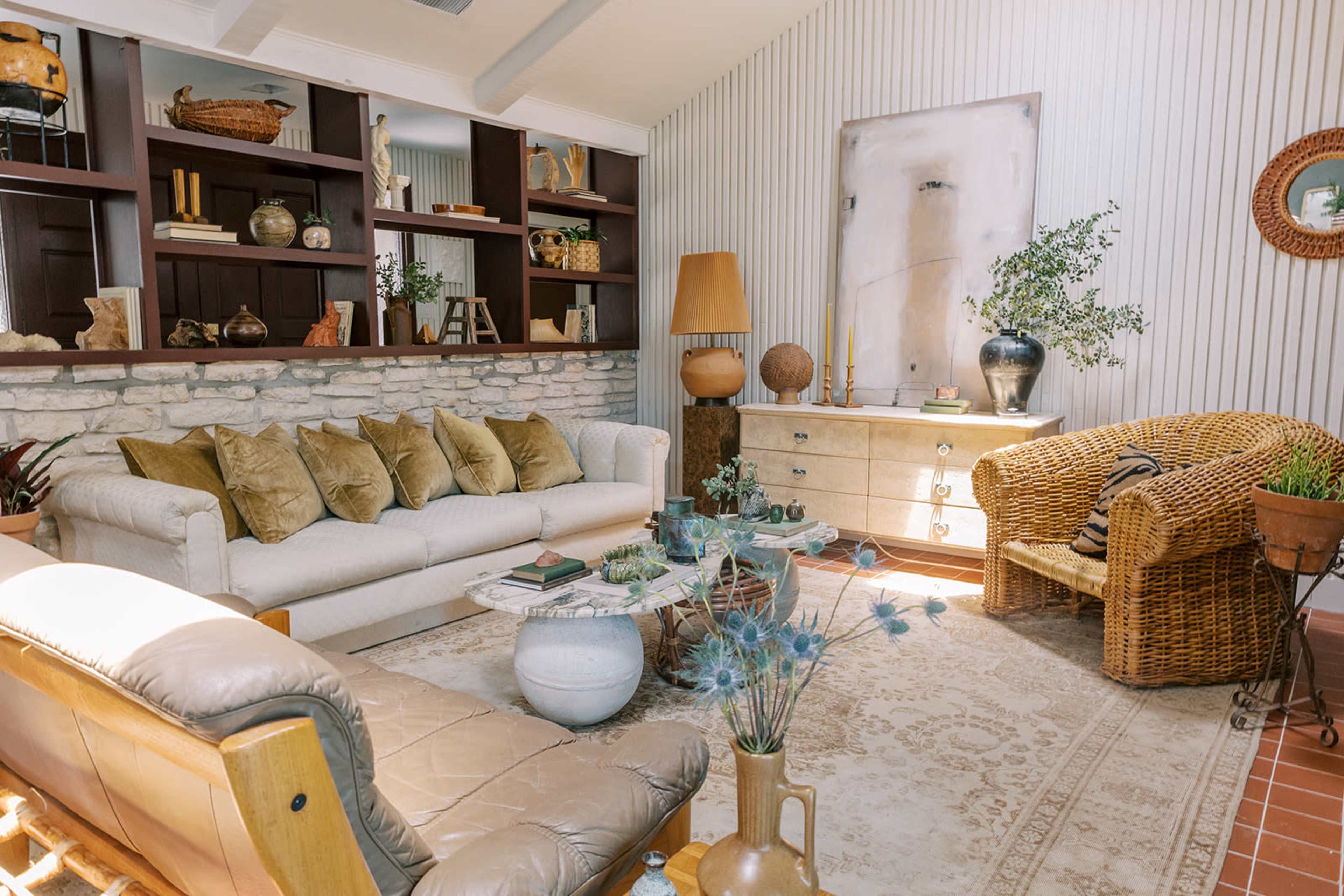 A cozy living room featuring a light-colored sofa with multiple cushions, a wicker chair, a wooden side table, and a stone wall bookshelf filled with decorative items.
