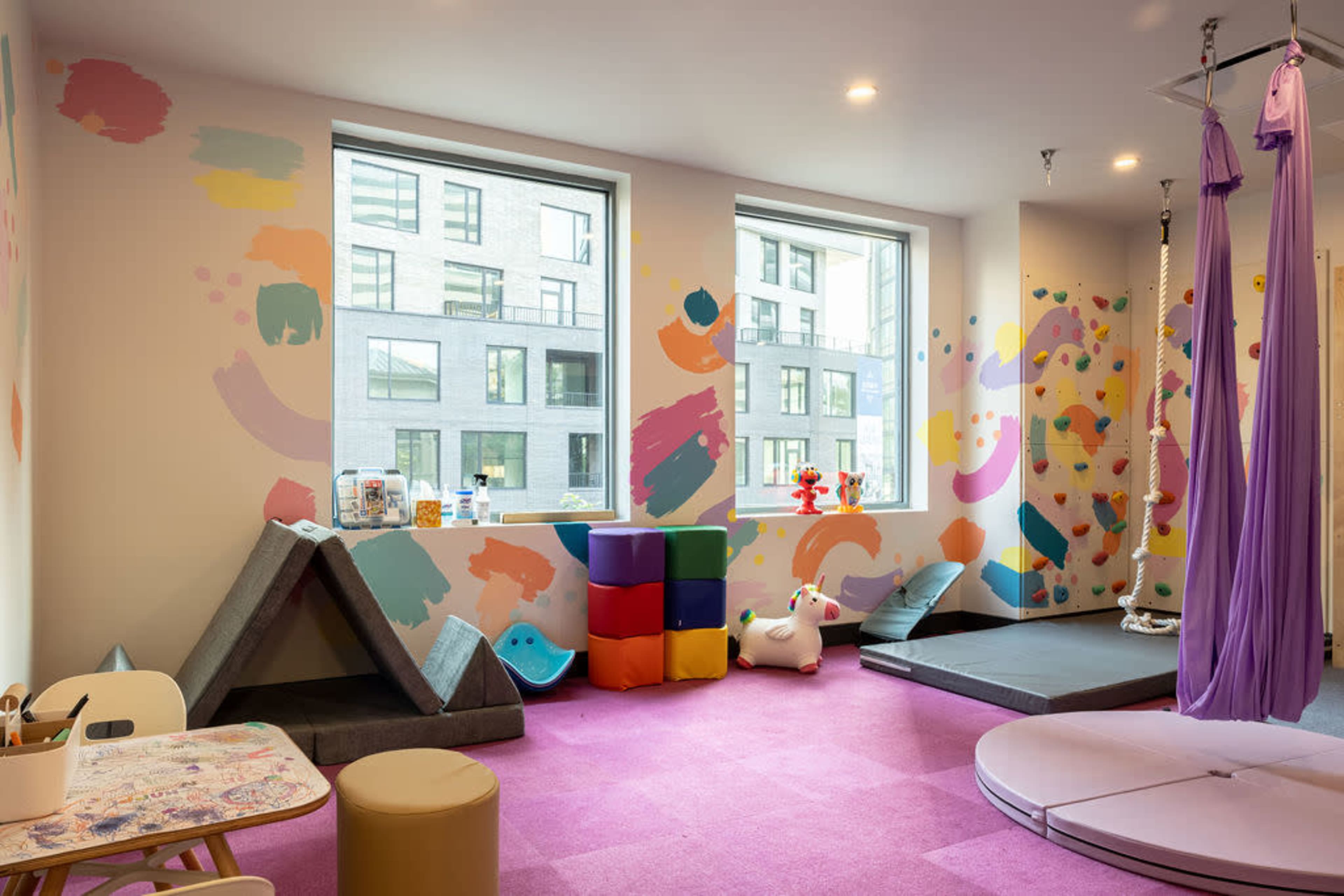 A brightly colored playroom features soft play equipment, hanging silks, and artwork on the walls.