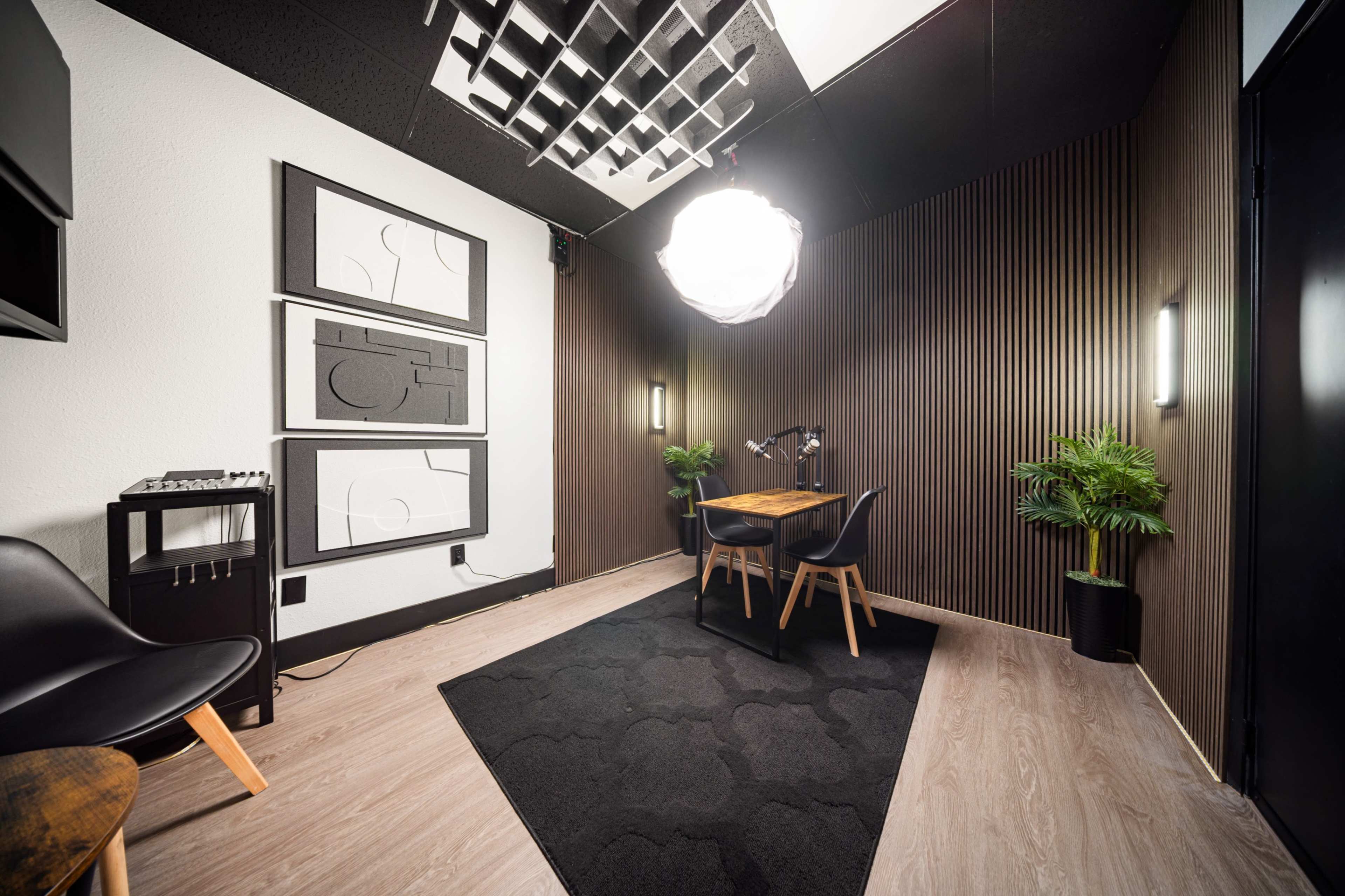 The image shows a modern, minimalist workspace featuring a wooden table and chairs, decorative wall art, and plants, set against a backdrop of textured wall panels and ceiling tiles.