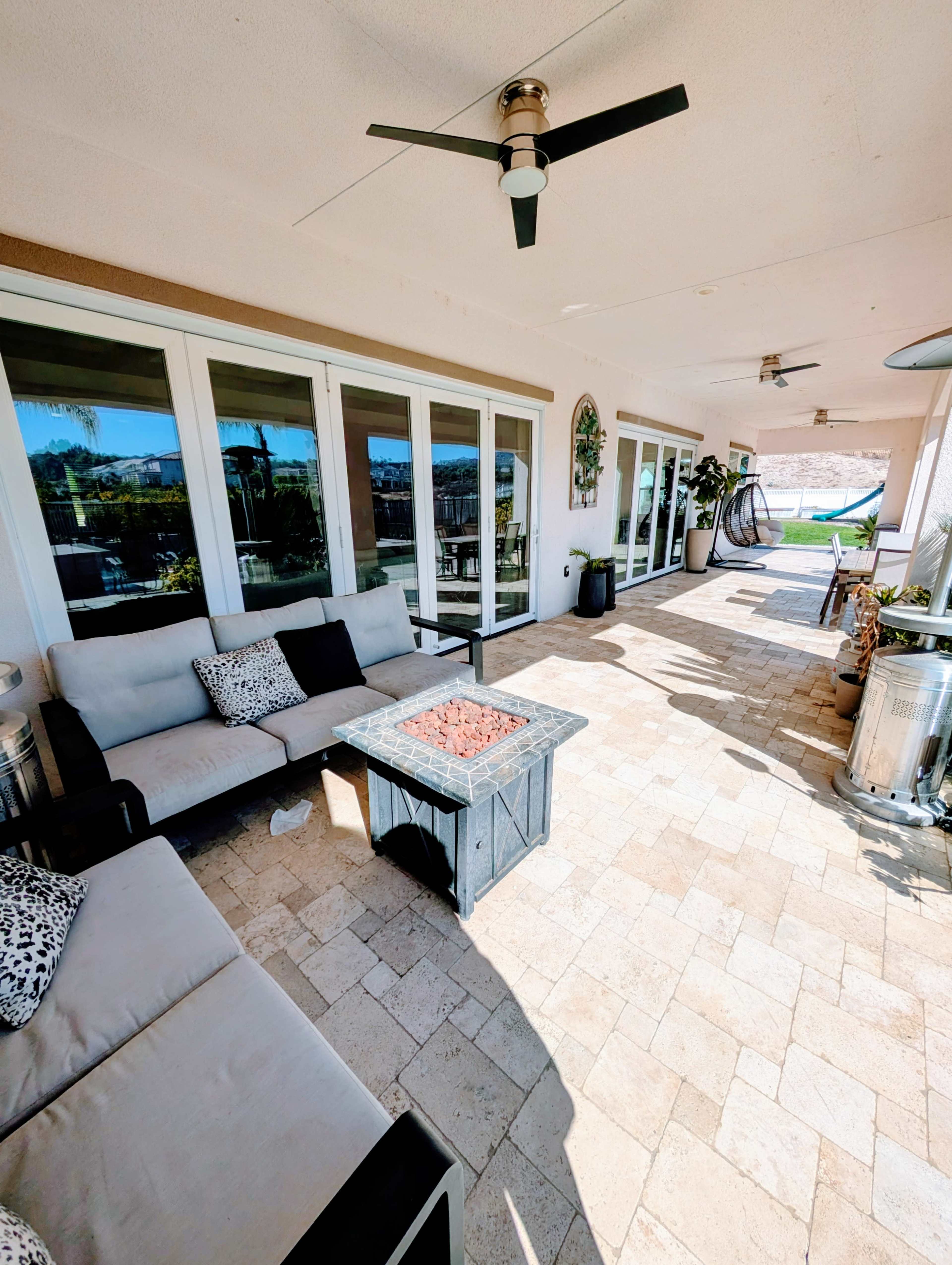 The image shows a spacious outdoor patio area with a seating arrangement, a fire pit, and large sliding glass doors leading into a house.