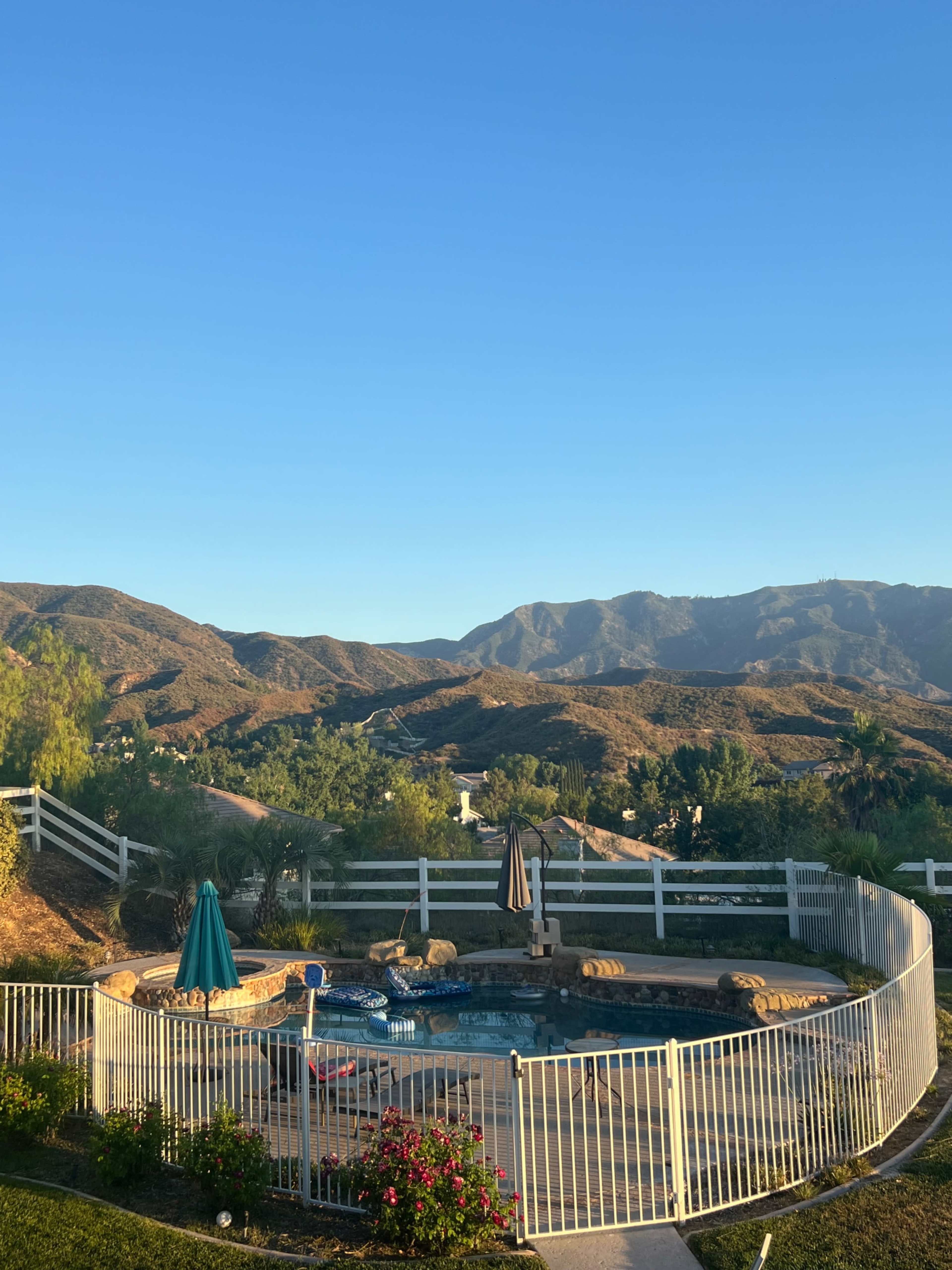 A fenced pool area is situated in a mountainous landscape under a clear blue sky.