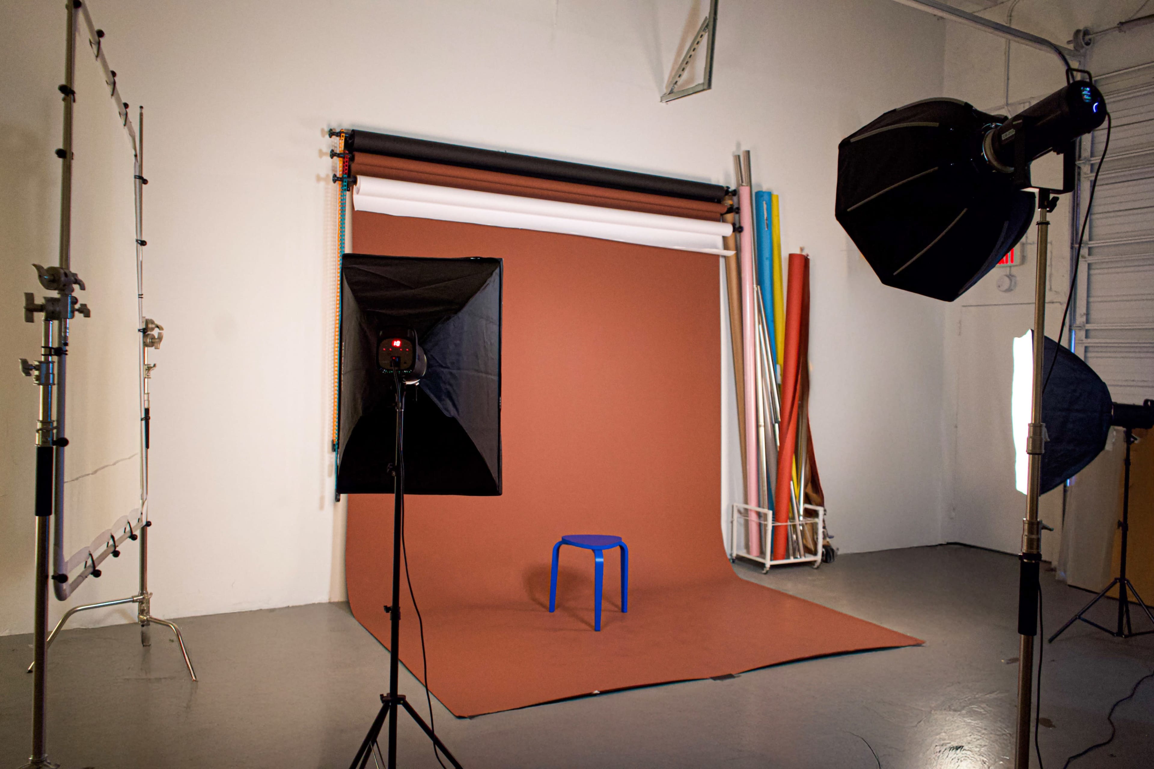 A photo studio setup features a brown backdrop, colorful rolls of paper, and various lighting equipment surrounding a blue stool.